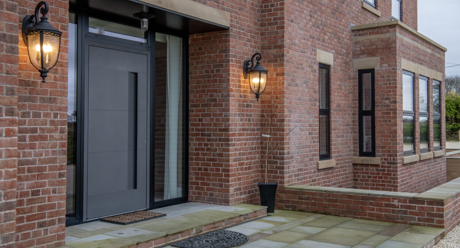 Modern brick house exterior with a sleek gray front door, two lit wall lanterns on either side of the entrance, large windows, and a paved stone walkway leading up to the front doors.