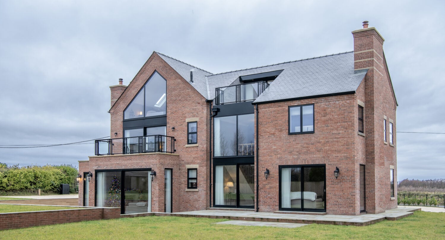 A modern three-story brick house with large glass windows and sliding doors, a rooftop balcony, and a sloped gray roof, set in a grassy yard with overcast skies in the background.
