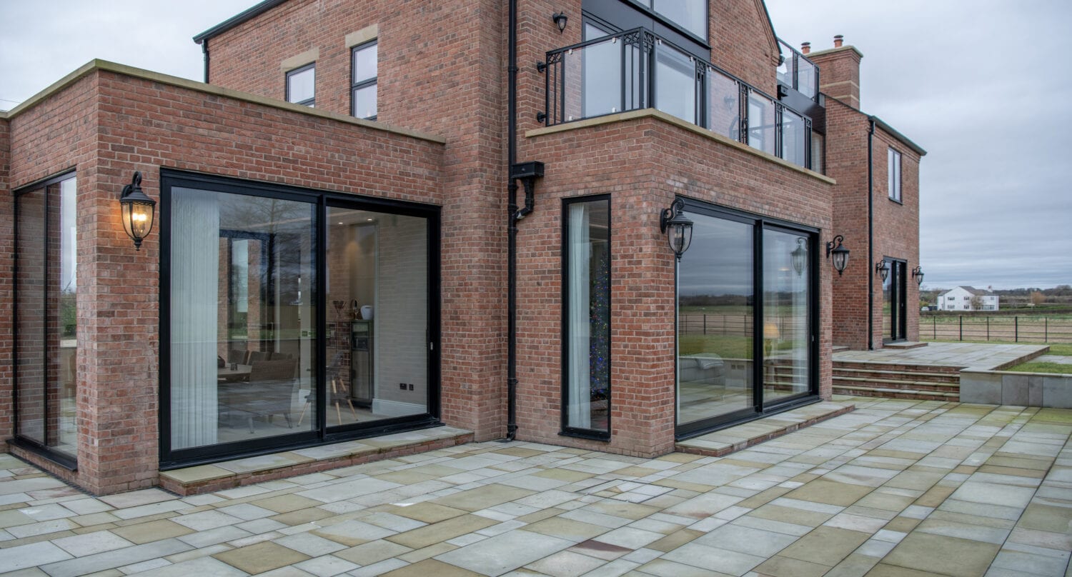 A modern red-brick house with large glass sliding doors and windows, sleek black trim, outdoor lanterns, and a spacious stone patio. The scene appears overcast with open fields in the background. Glass roofing adds a contemporary touch.