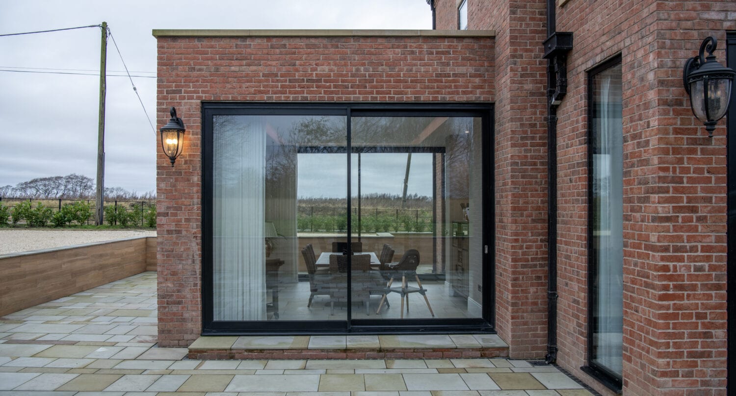 A modern brick house with large sliding doors opening to a patio area. Indoor dining table and chairs are visible through the glass. Outdoor wall-mounted lantern lights and tiled ground complete this bespoke glass solution.