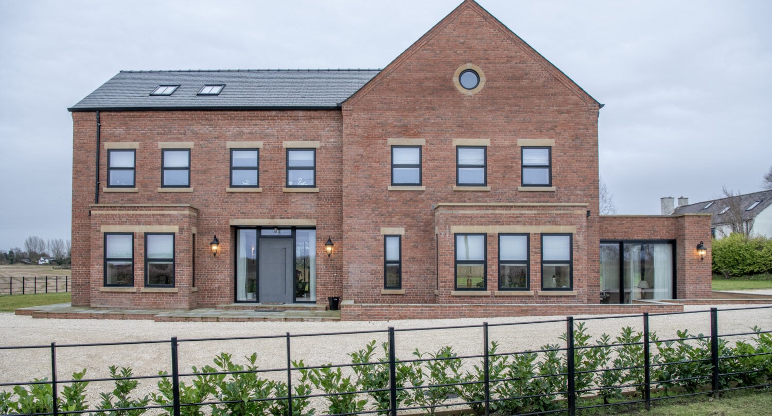 A large, modern red-brick house with black-framed windows, bifold doors, a slate roof, and a round window near the peak. The property is surrounded by a gravel driveway, plants, and a black metal fence.