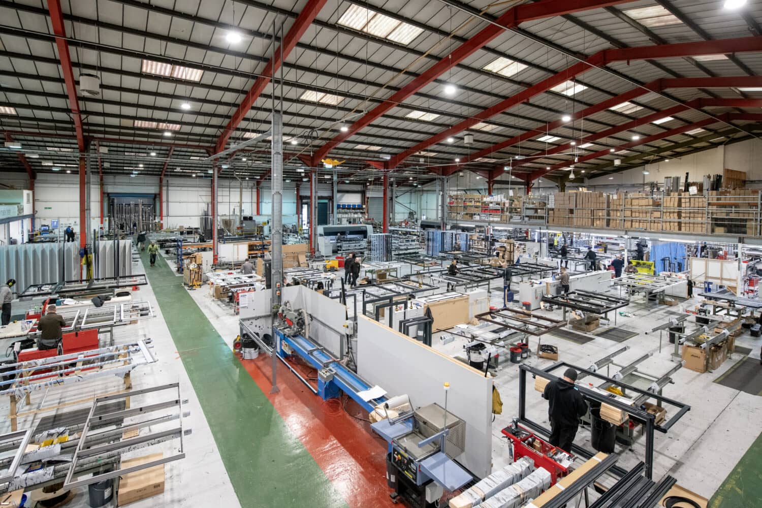 A large, brightly lit factory interior with high ceilings and red steel beams features various workstations where workers assemble metal front doors and bespoke glass solutions. Shelves of materials and machinery are visible throughout the organized space.