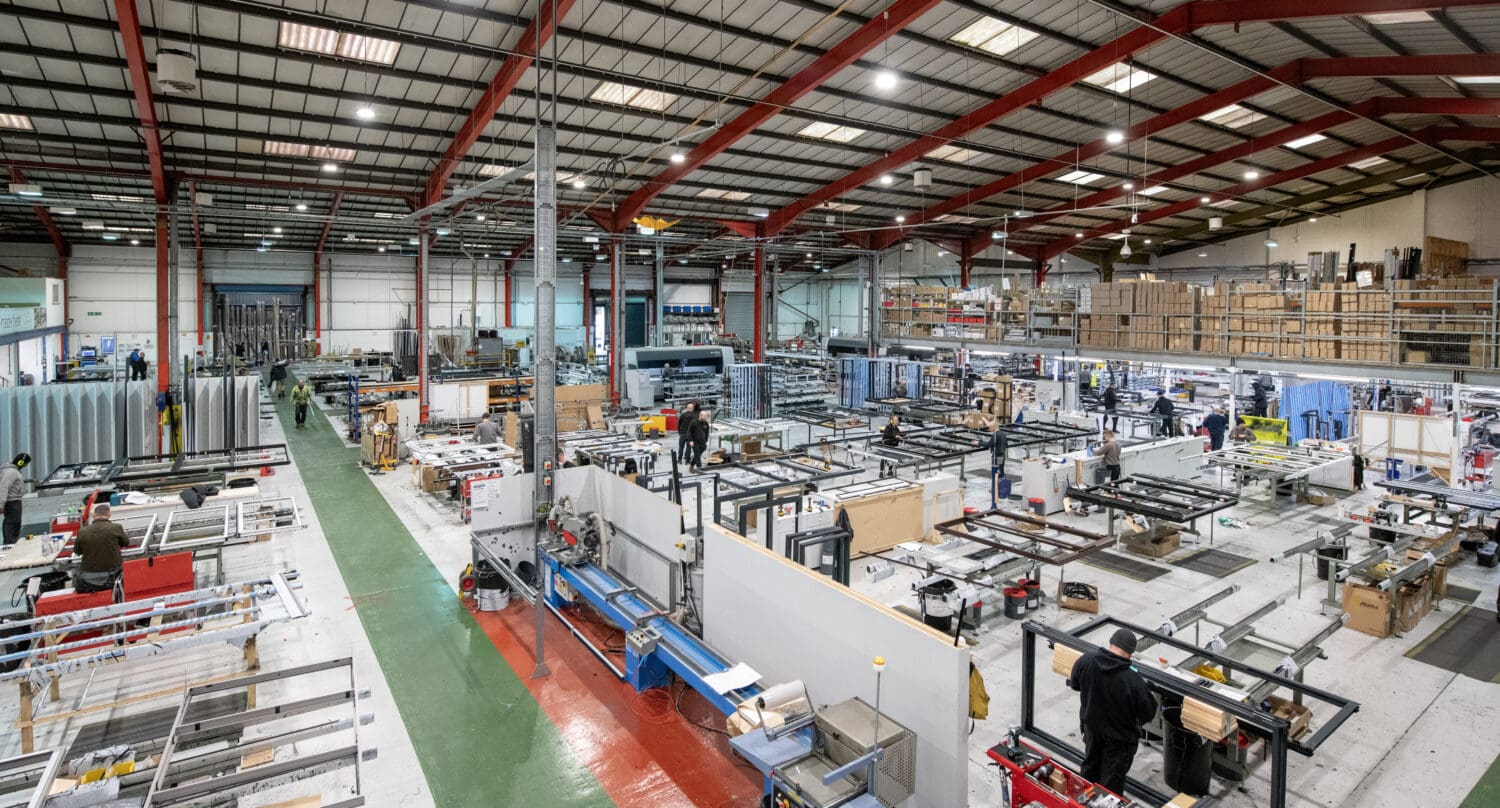 A large, brightly lit factory interior with high ceilings and red steel beams features various workstations where workers assemble metal front doors and bespoke glass solutions. Shelves of materials and machinery are visible throughout the organized space.