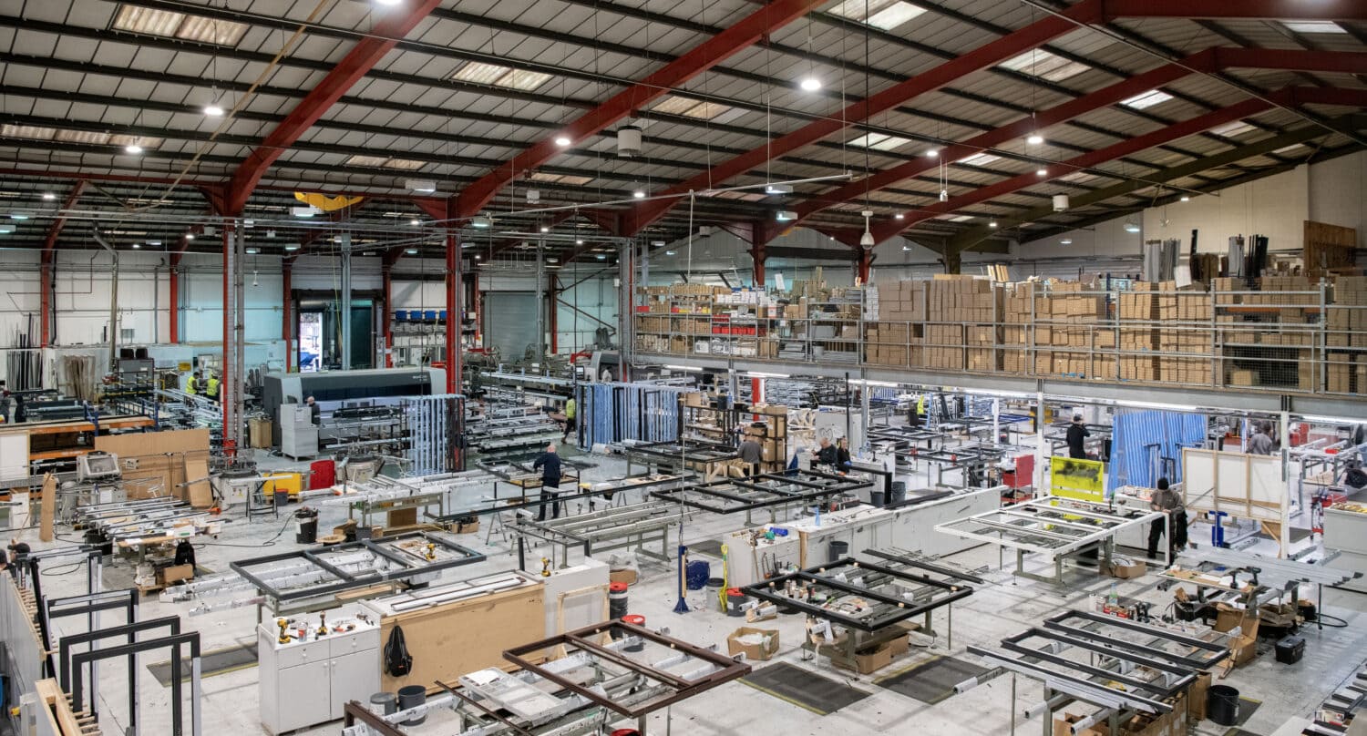 A large, organized factory floor with high ceilings, metal beams, and skylights. Various machinery and workstations fill the space, with workers engaged in assembly tasks near industrial bifold doors that allow easy movement of materials.