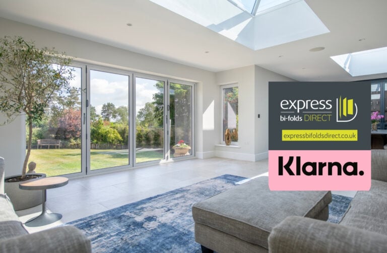 Bright, modern living room with large glass doors opening to a garden, skylights, a blue rug, two sofas, and an indoor plant. Bespoke glass solutions by Express Bi-Folds Direct and Klarna payment logos are displayed.