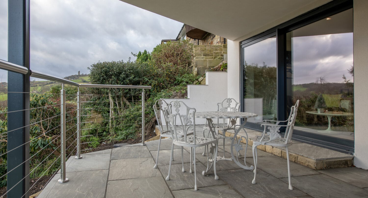 A covered patio with a white metal table and four matching chairs on stone tiles, overlooking a green hillside. Sliding doors lead inside, and there are steps and a metal railing on the left.