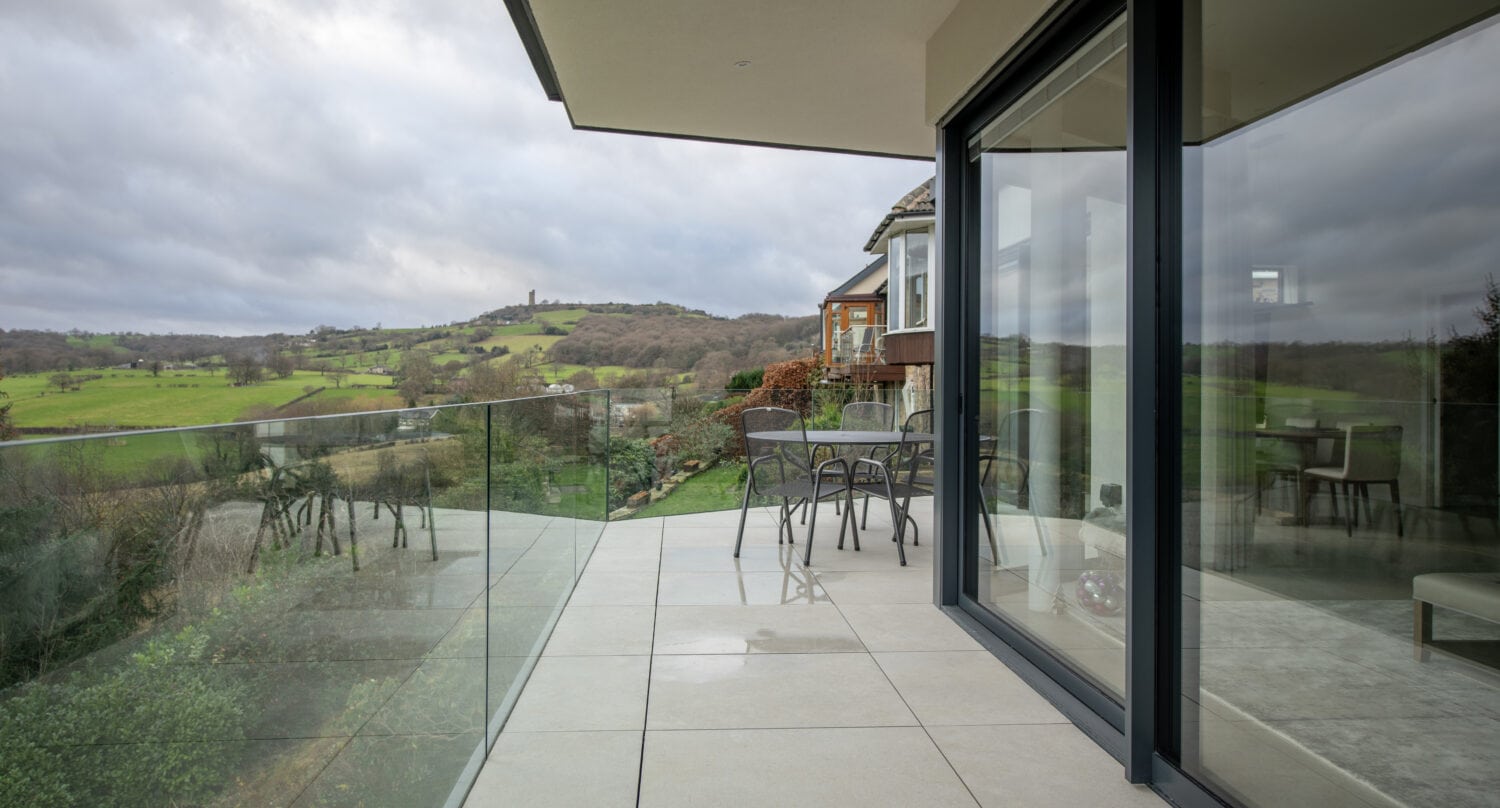 A modern balcony with glass railing and sliding doors overlooks a green countryside landscape. There is a small table with chairs, and large glass doors connect the balcony to the interior. Rolling hills and cloudy skies are visible in the background.