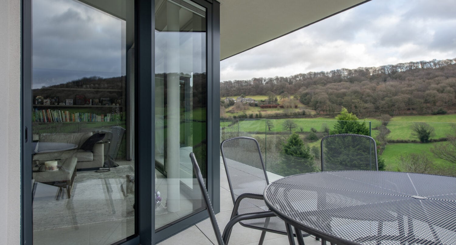 A sliding glass patio door opens to a balcony with a metal table and chairs, overlooking scenic green hills, trees, and cloudy skies. Indoors, a bookshelf and armchair are visible for cozy relaxation.