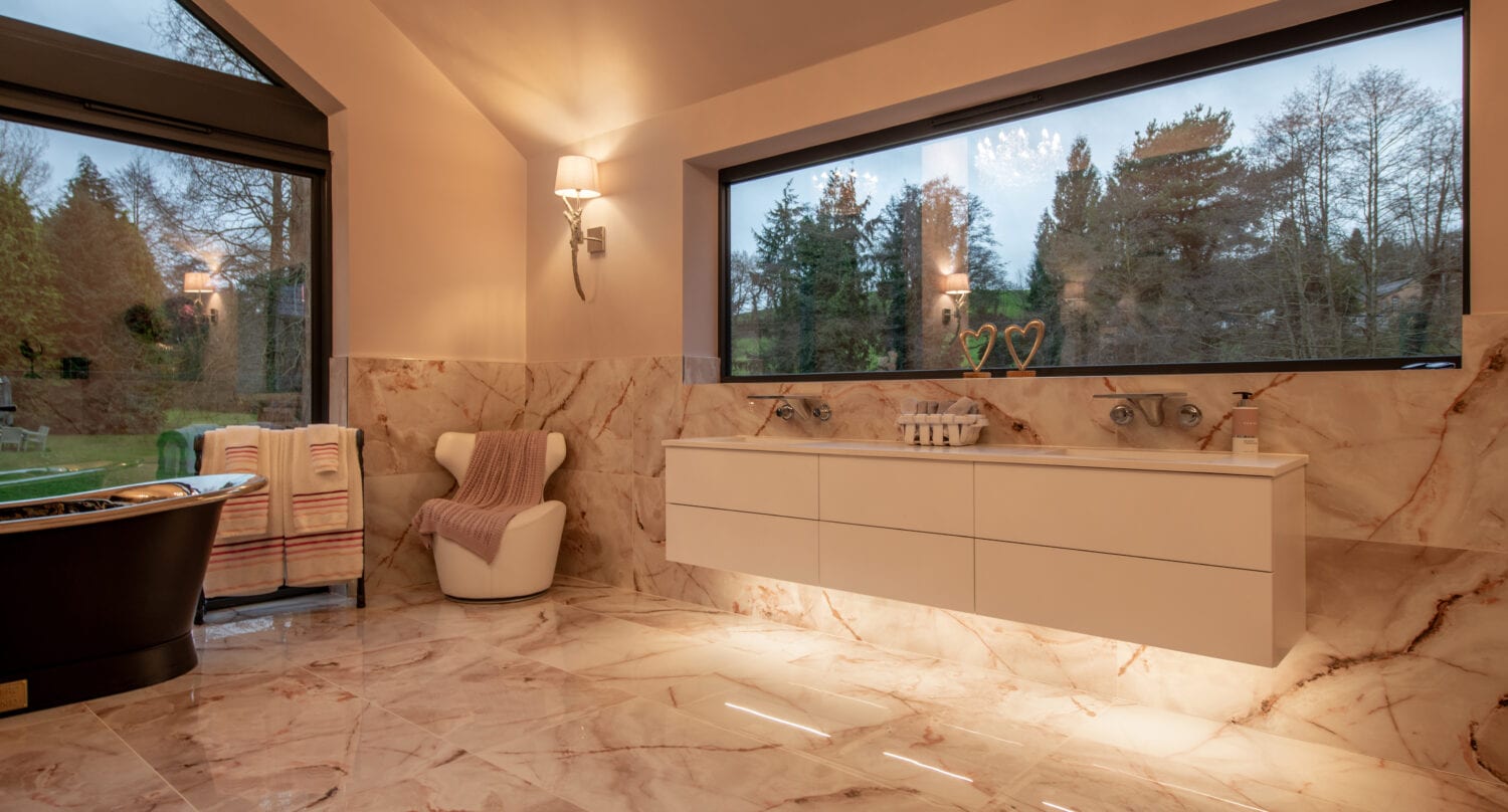 Luxurious bathroom with marble floors and walls, a large window overlooking trees, modern floating double sink, black tub, and a cozy chair with towels. Bespoke glass solutions and warm lighting create an inviting atmosphere.