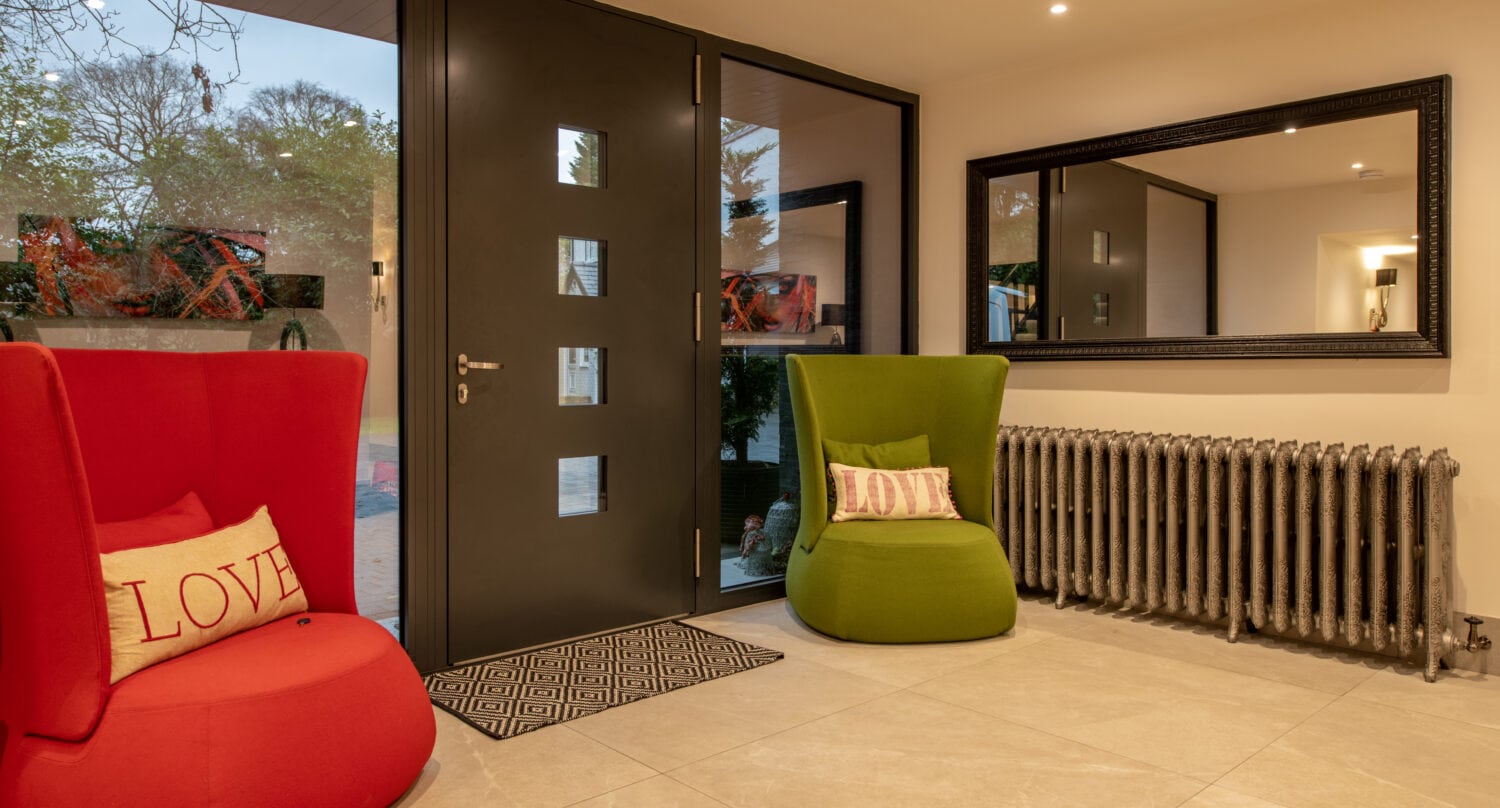 Modern entryway with large gray front doors, red and green armchairs each with a LOVE pillow, a geometric doormat, large wall mirror, and a metal radiator. Trees and garden visible through the glass panels.