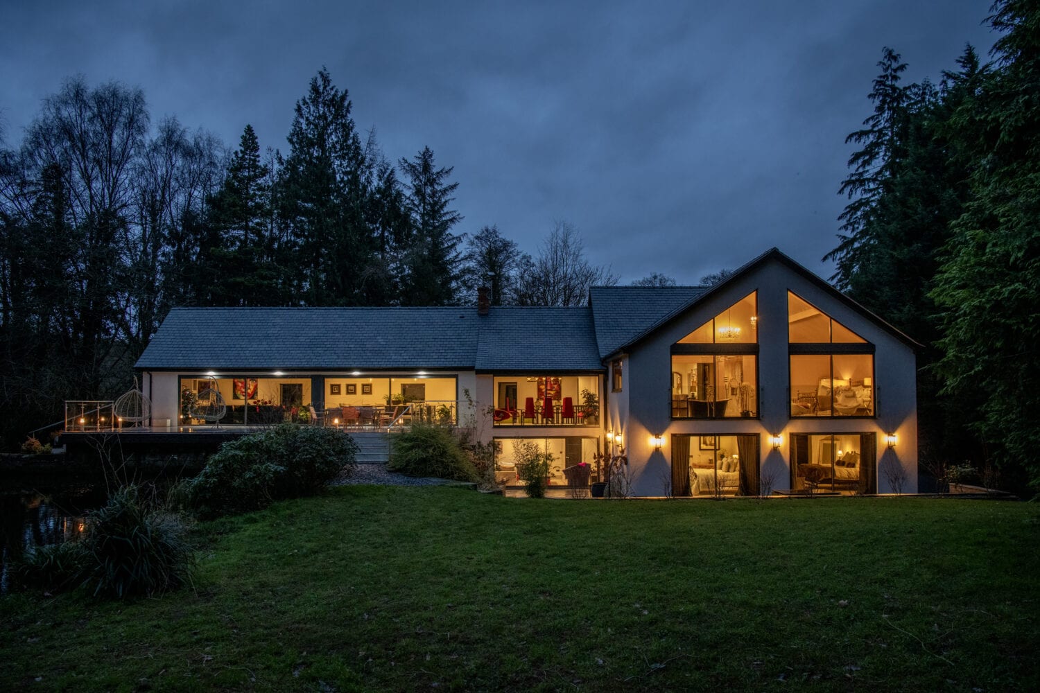 A modern house with large windows and sliding doors glows warmly at dusk, surrounded by trees and a grassy lawn. The lights inside highlight cozy interior spaces, creating a welcoming atmosphere against the evening sky.