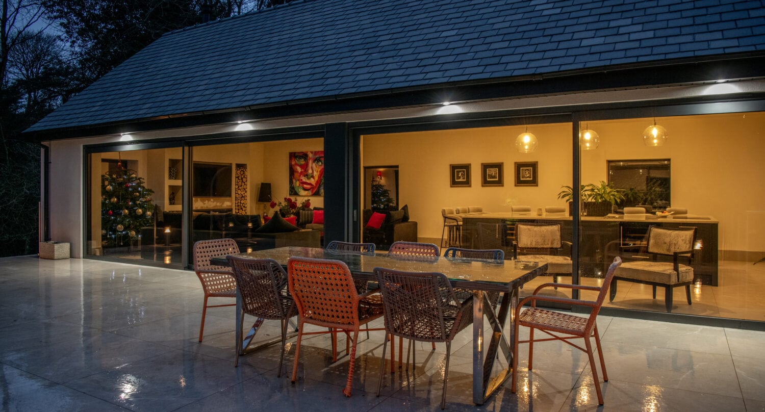 Modern patio at dusk with a glass dining table and colorful chairs on a wet tiled floor. Warm light fills the living area inside, where a Christmas tree and art are visible through bespoke glass solutions and large sliding doors.