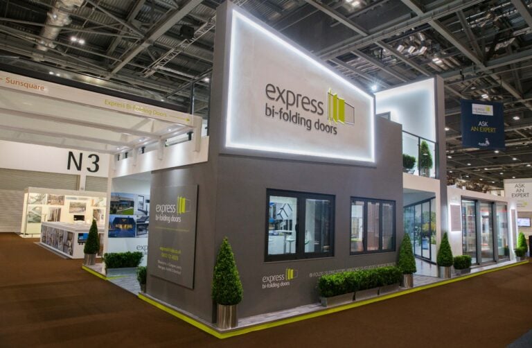 A modern exhibition booth for Express Bi-Folding Doors, featuring large glass windows, bespoke glass solutions, branded signage, potted plants, and a sleek gray and white design, set inside a spacious convention center.