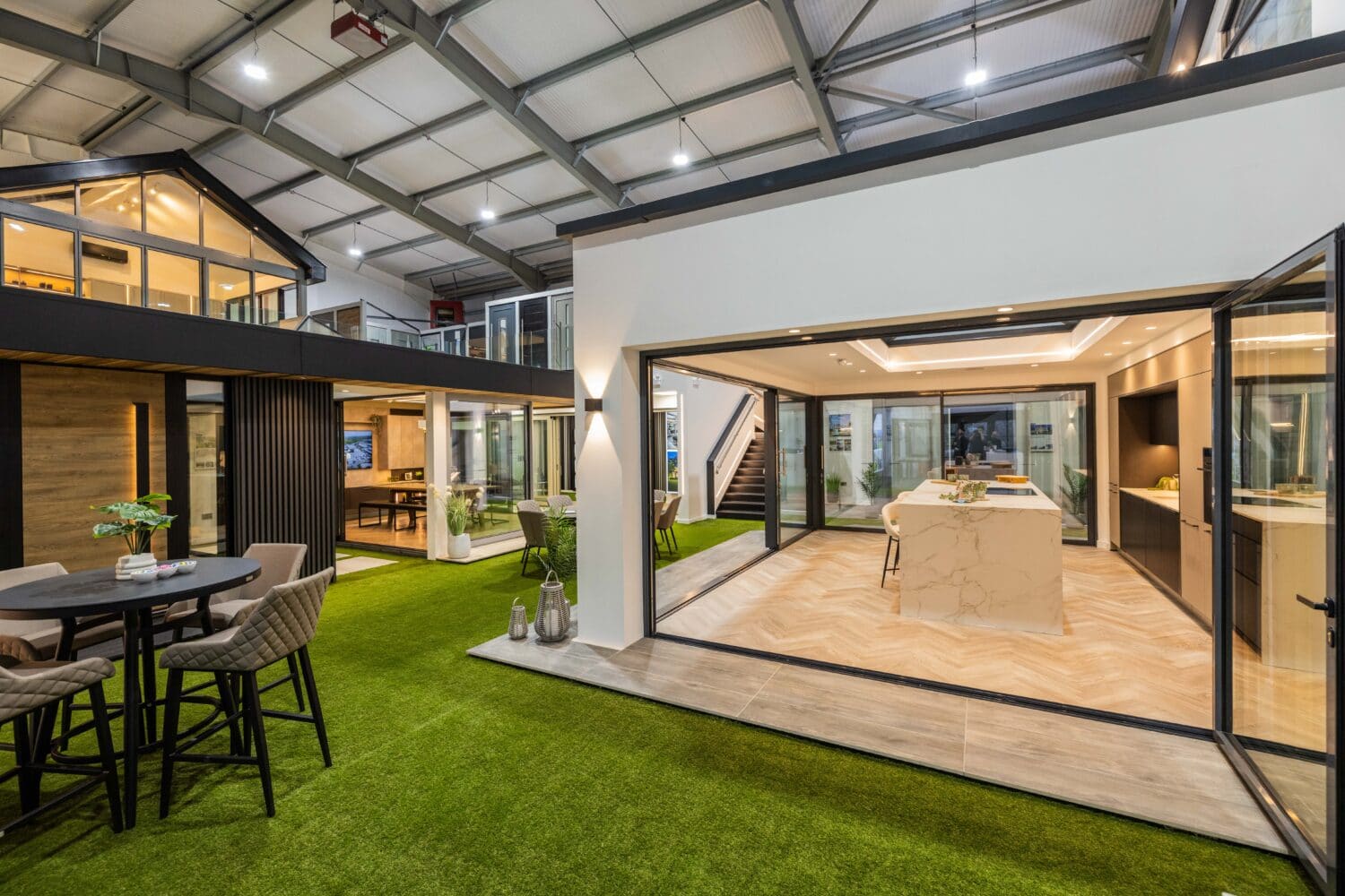 Modern indoor showroom with model homes, open kitchen, dining area, artificial grass flooring, tables with chairs, expansive bifold doors, large windows, and bright lighting under a high industrial ceiling.