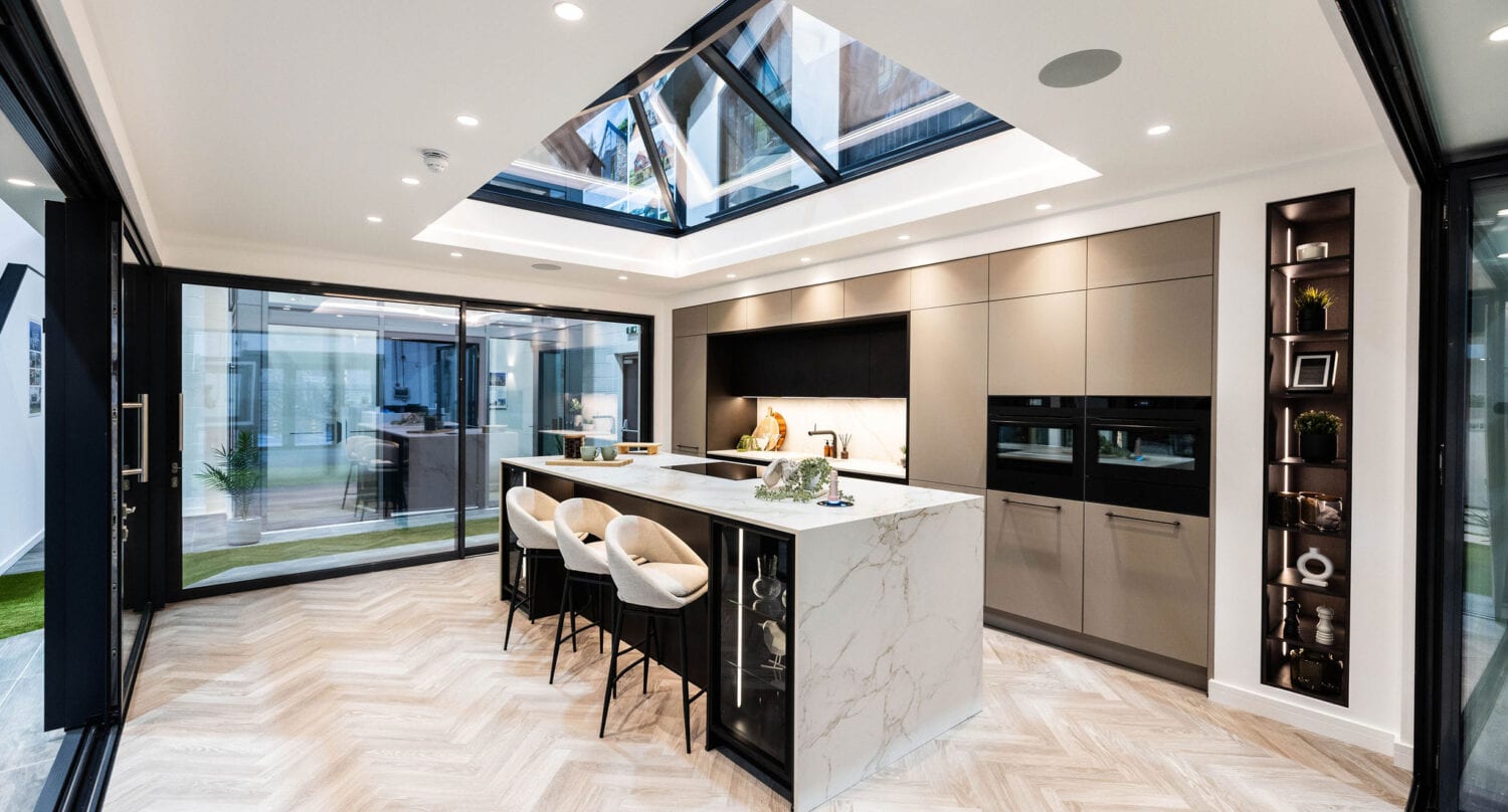 Modern kitchen with marble island, three white stools, sleek cabinetry, built-in ovens, and a large skylight above. Bifold doors and ample lighting create a bright, open atmosphere with a view of the outdoors.