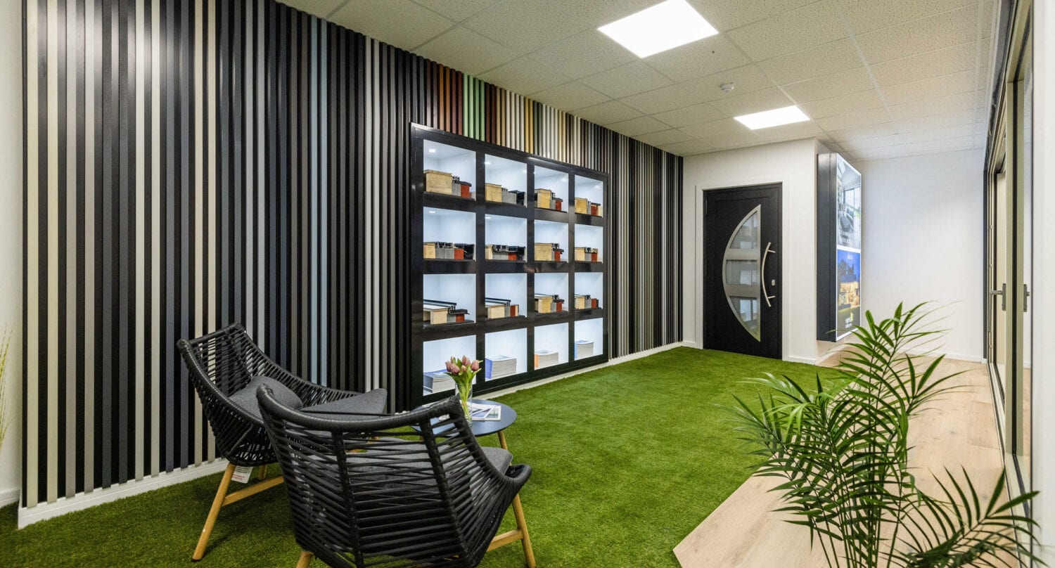 Modern office lounge area with artificial grass flooring, two black woven chairs around a small round table, striped accent wall with shelves displaying samples, potted plants, and natural light streaming in through sleek bifold doors.