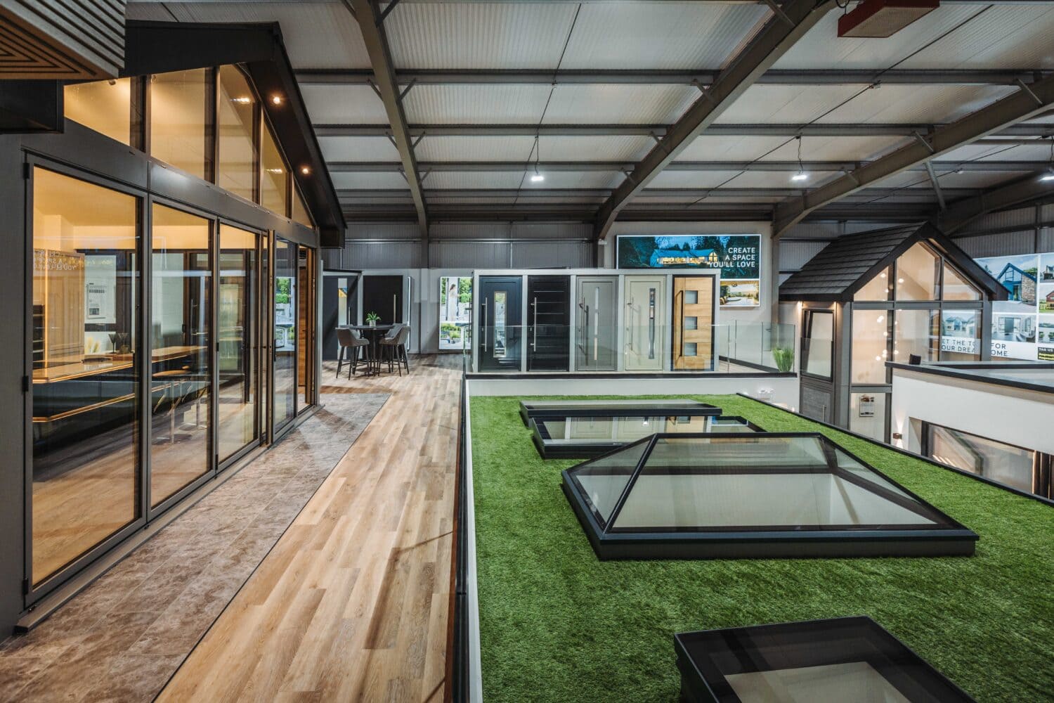 A modern indoor showroom displays large glass windows, sliding doors, and skylights. The space features wood and carpet flooring, artificial grass, and contemporary architectural elements under a metal roof.