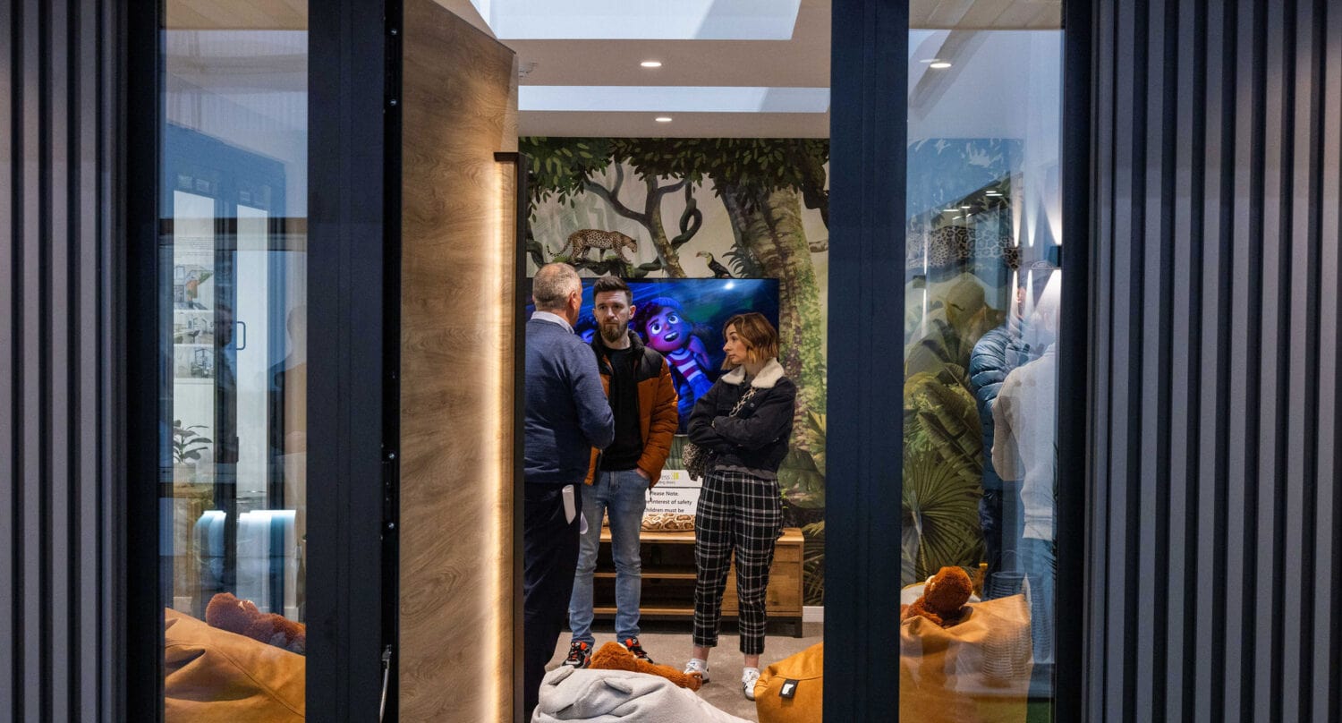 A group of people stand and talk inside a cozy, modern room with bean bags, large windows, and a jungle-themed mural. Nearby, sleek sliding doors open to another area, while a bright TV screen displays an animated character in the background.