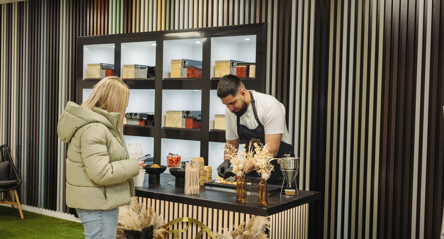 A man in an apron stands behind a counter preparing food while a woman in a green jacket waits. Behind him, shelves with boxes and jars sit beneath striking glass roofing, and dried floral decor accents the striped paneled wall.
