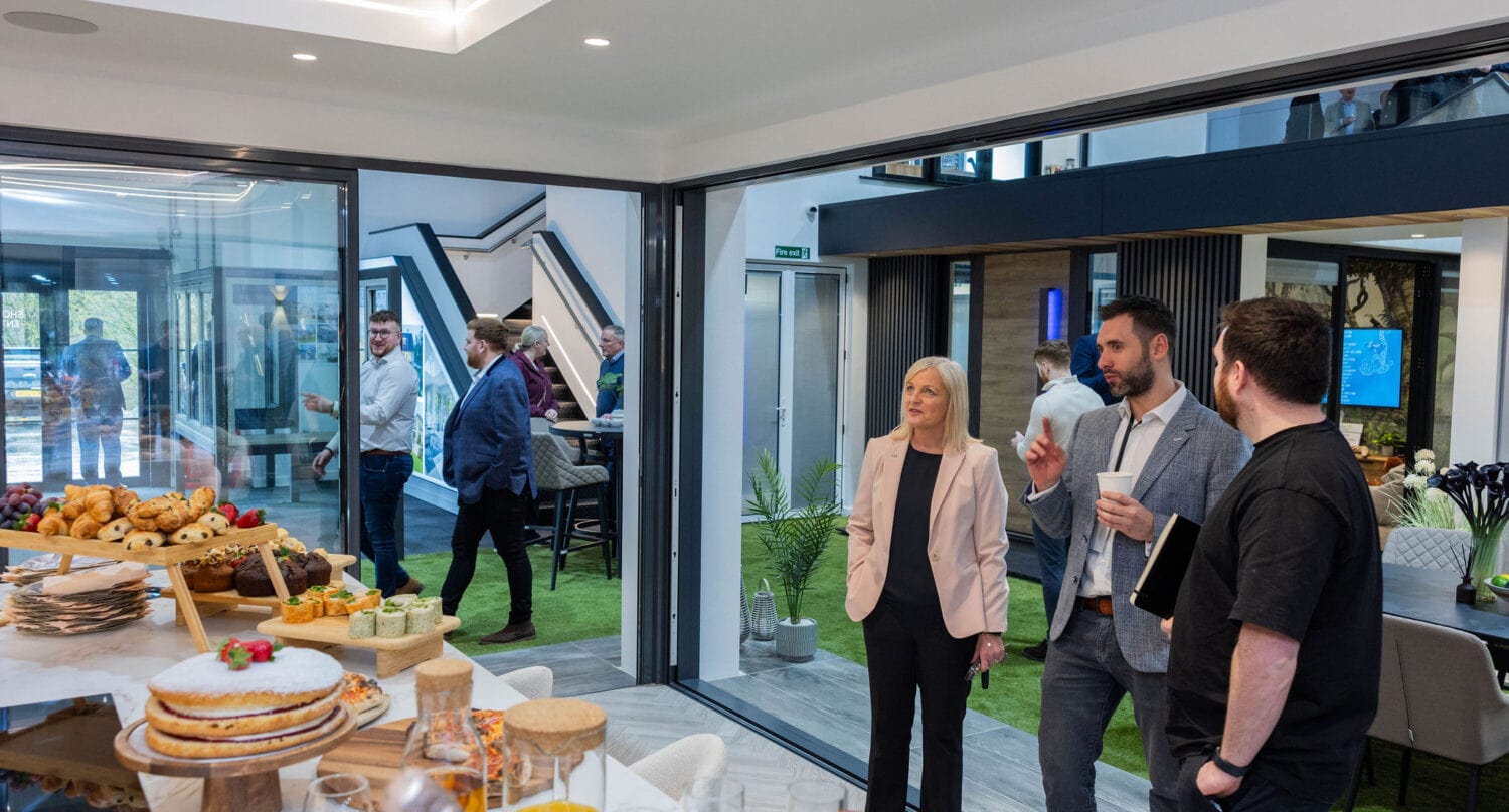 A group of people in business attire converse in a modern office with bespoke glass solutions and sliding doors. In the foreground, a table is set with cakes, pastries, and drinks as others mingle near large windows.
