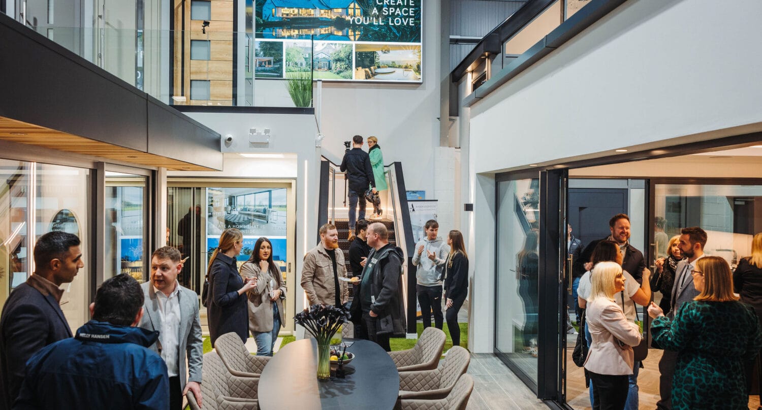 A group of people socialize and network inside a modern showroom with large windows, sliding doors, a central table with refreshments, and a staircase leading to a mezzanine level.