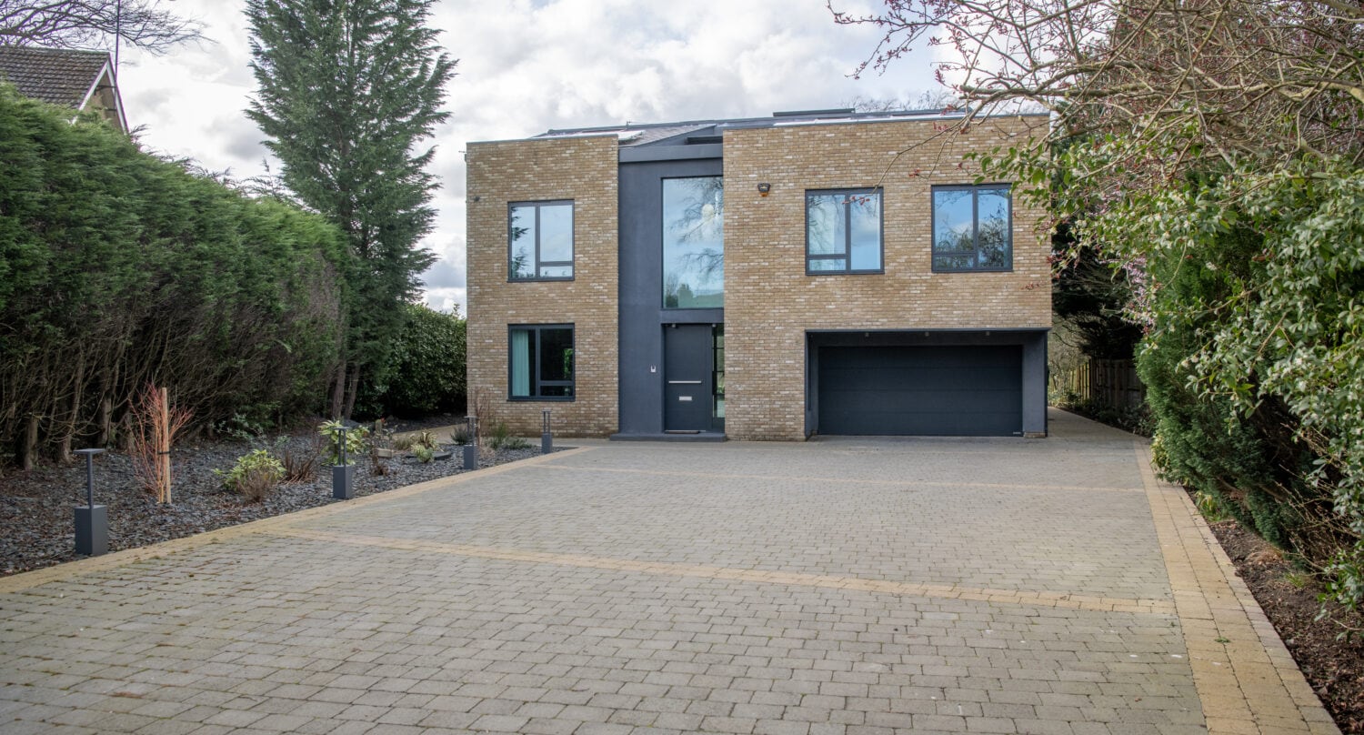 A modern two-story house with a brick exterior, large windows, bespoke glass solutions, and a black garage door, set back from the street with a spacious paved driveway and landscaping on both sides.