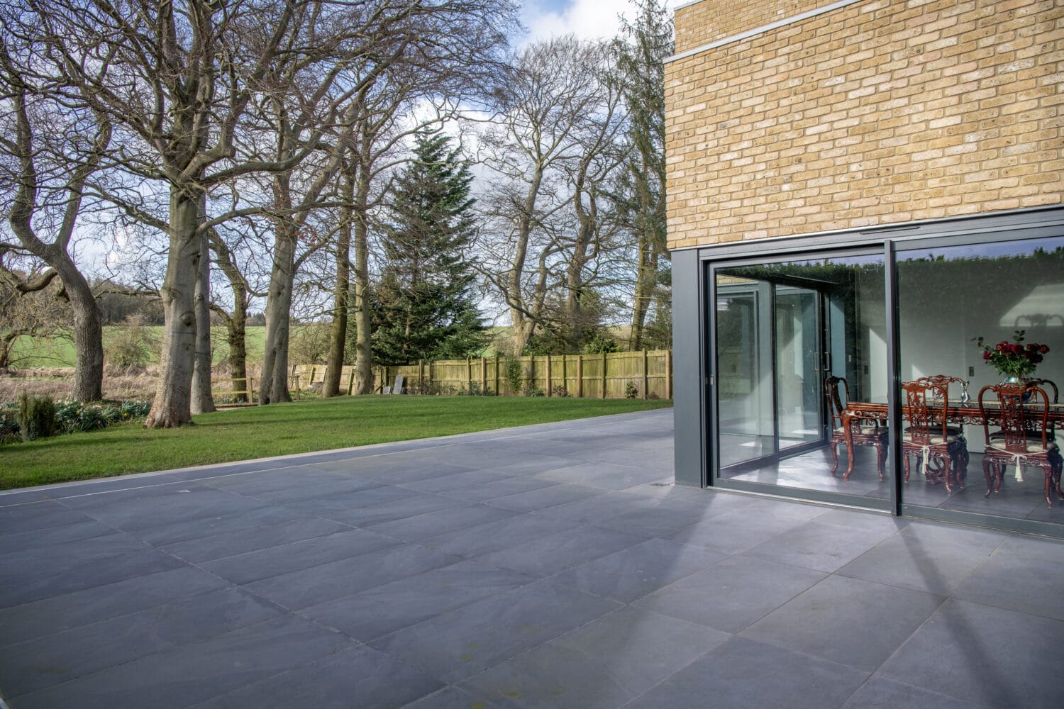 Modern house with bespoke glass solutions, featuring large sliding doors opening onto a spacious gray tiled patio that overlooks a grassy yard with leafless trees and a wooden fence. A dining table with chairs is visible inside.