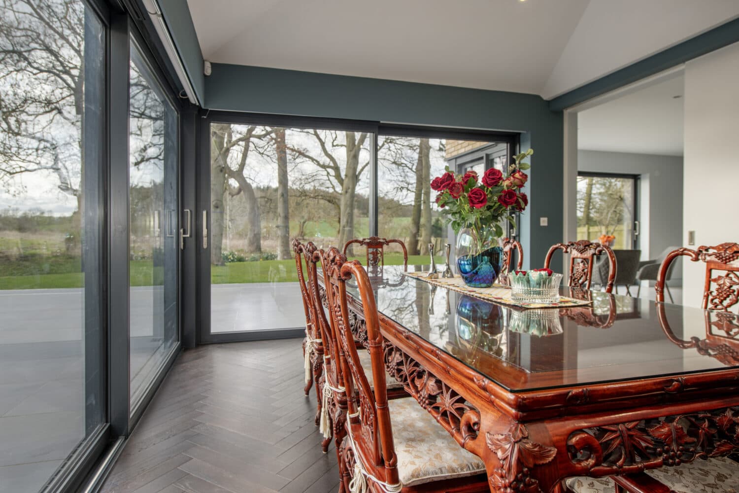 A dining room with carved wooden chairs around a polished table, a vase of red roses as centerpiece, and large bifold doors offering a view of trees and greenery outside.