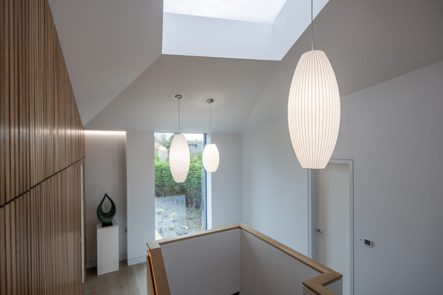 Modern interior hallway with wooden slat wall, skylight, two large white pendant lights, a staircase with wooden railing, sliding doors opening to greenery, and a white sculpture on a pedestal.