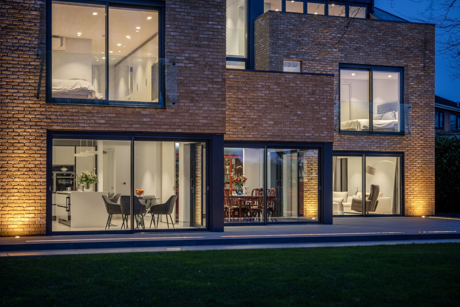 Modern brick house at dusk, with large glass windows and stylish bifold doors revealing well-lit interior spaces including dining and living areas. Bedrooms on the upper floor and exterior lights illuminate the facade.