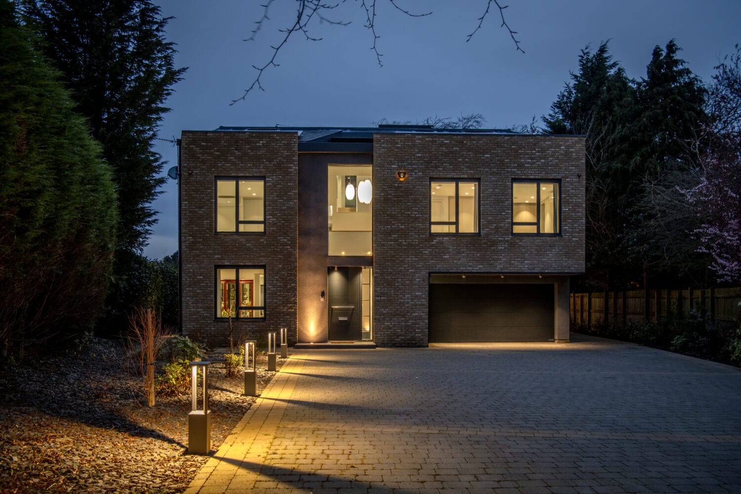 Modern two-story house with large windows, brick exterior, and a double garage, featuring bespoke glass solutions like sliding doors. Warm outdoor lights illuminate a paved driveway at dusk, surrounded by trees and landscaped garden beds.