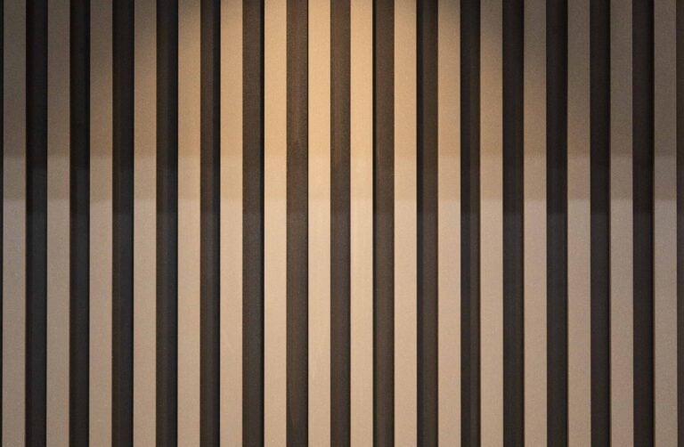 Vertical wooden slats on a wall are illuminated by a single recessed ceiling light, creating alternating shadows and highlights. The ceiling features wooden trim, complementing the modern look often found with sliding doors.
