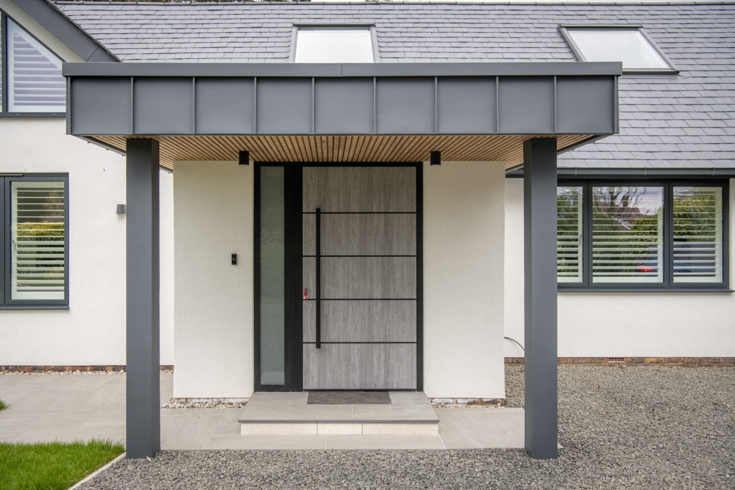 Modern house entrance with a wide, light wood front door, large sidelights, and a gray overhang supported by two pillars. White walls and horizontal windows with shutters frame the front doors, while gravel and grass complete the inviting look.