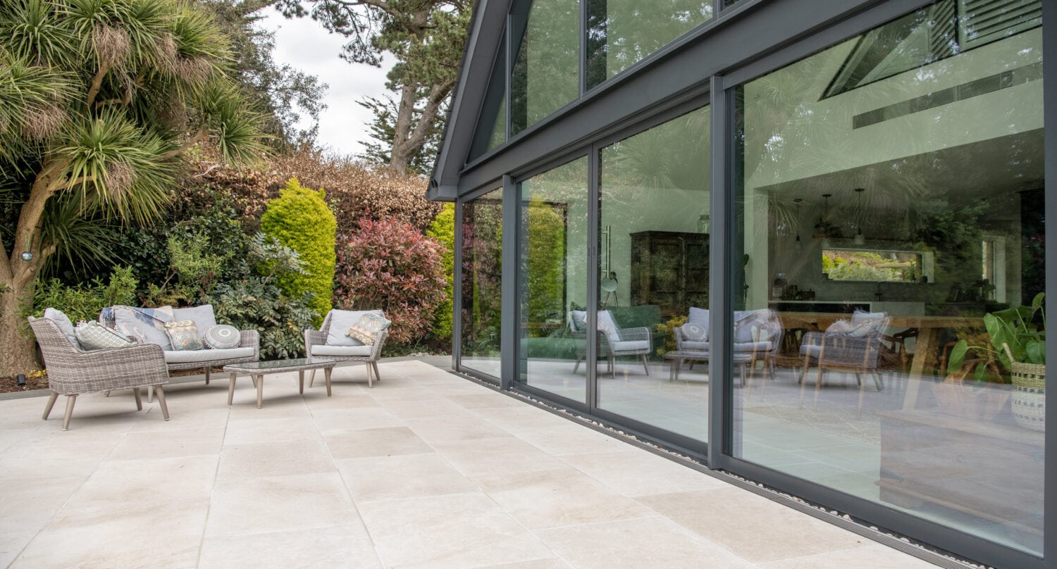 A modern patio with light stone tiles features outdoor seating and is bordered by lush greenery. Bespoke glass solutions, including large sliding doors, seamlessly connect the patio to a stylish, open-plan indoor living space.