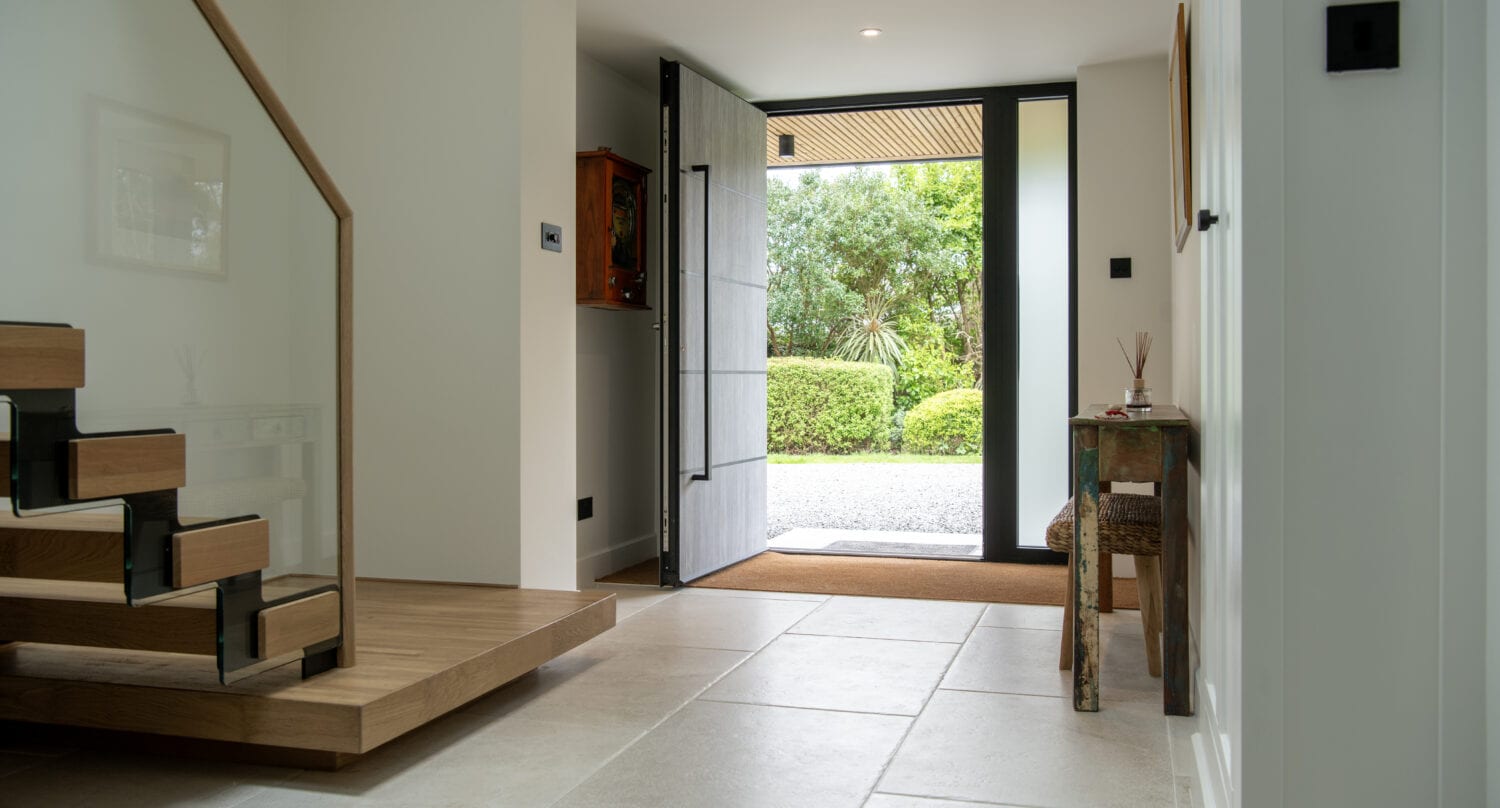A modern, bright entryway with large floor tiles, an open wooden staircase with bespoke glass solutions for the railing, a wooden console table, and a wide front door open to a green garden outside.