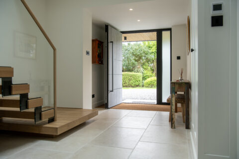 A modern, bright entryway with large floor tiles, an open wooden staircase with bespoke glass solutions for the railing, a wooden console table, and a wide front door open to a green garden outside.