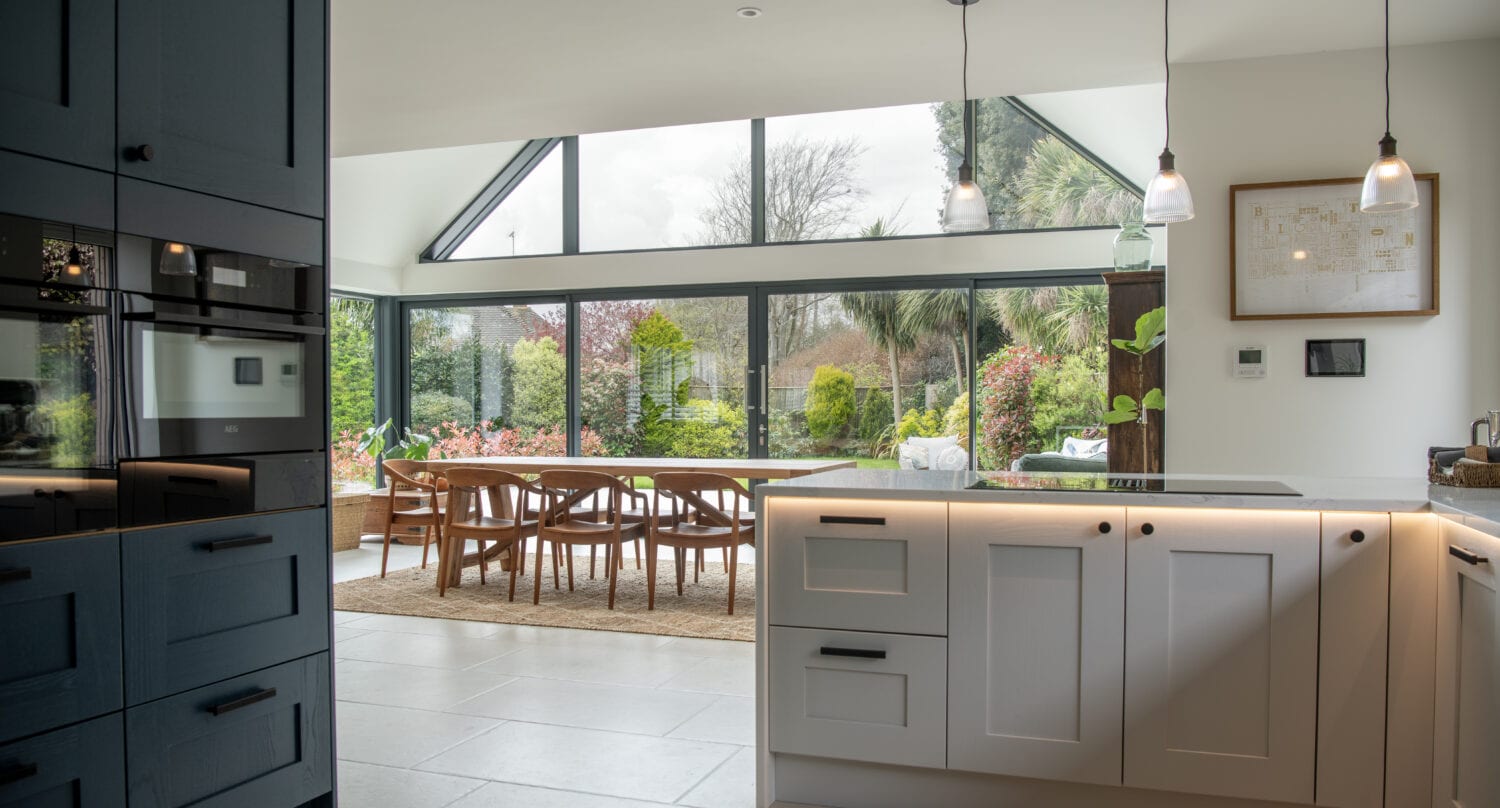 Modern kitchen with light cabinets and an island, leading to a dining area with wooden chairs and a large table. Floor-to-ceiling bifold doors showcase a lush garden outside. Pendant lights hang from the ceiling.