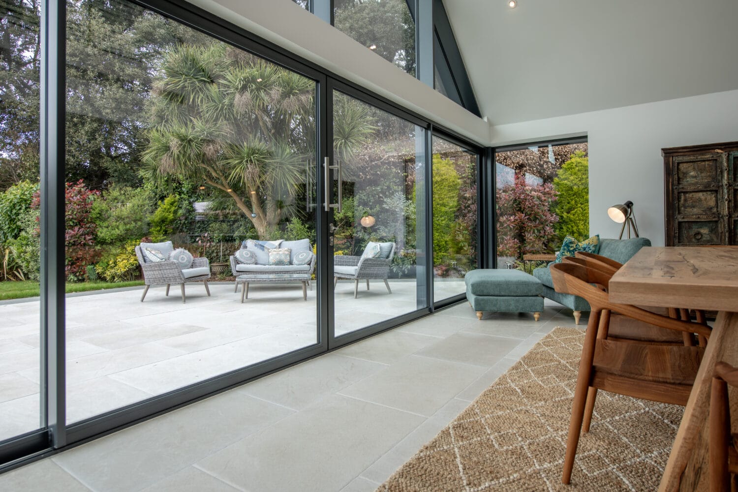 Spacious modern living area with large bifold doors opening to a patio with outdoor seating, surrounded by lush green garden trees and plants; indoor area has wooden dining table, chairs, and a cozy armchair.