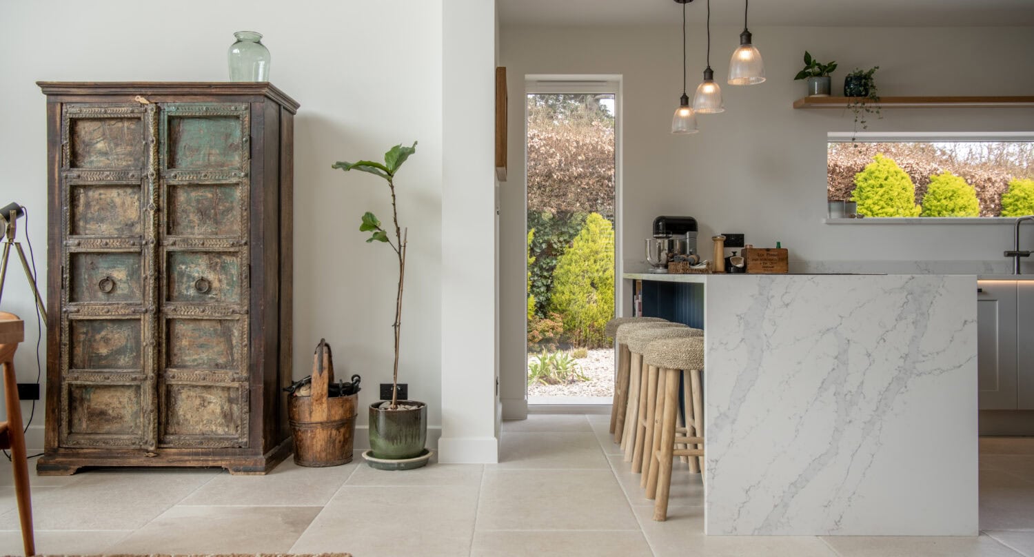 A bright, modern kitchen with a marble island, wooden stools, hanging pendant lights, a rustic cabinet, potted plants, and bespoke glass solutions like sliding doors leading to a sunny garden with green hedges.