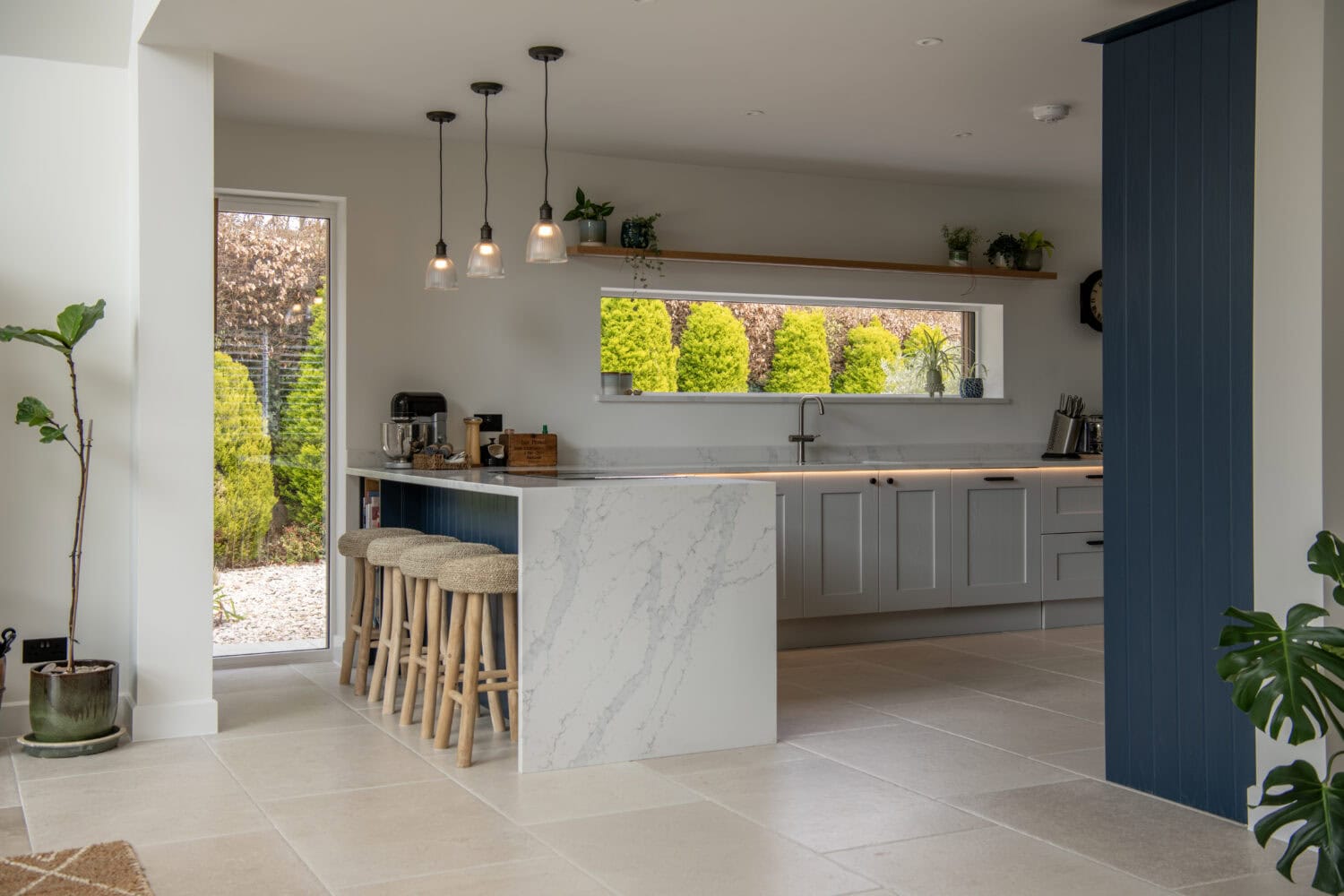 Modern kitchen with a marble island, three wooden bar stools, pendant lights, light gray cabinets, and large windows featuring bespoke glass solutions that frame views of green shrubs and a small garden outside.