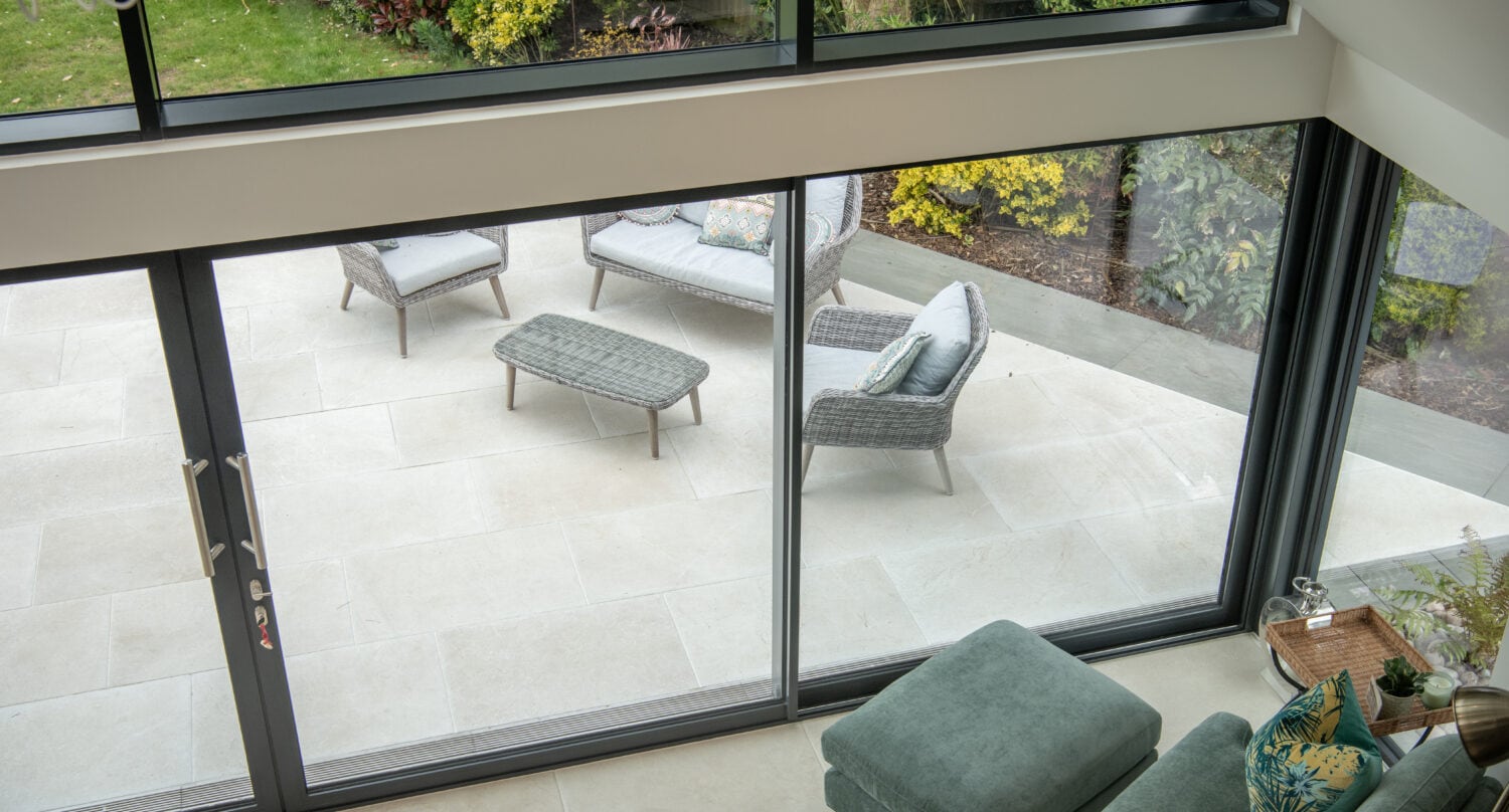 View from inside a modern home, looking through large bifold doors onto a patio with wicker furniture, surrounded by greenery and flowering plants in a garden.