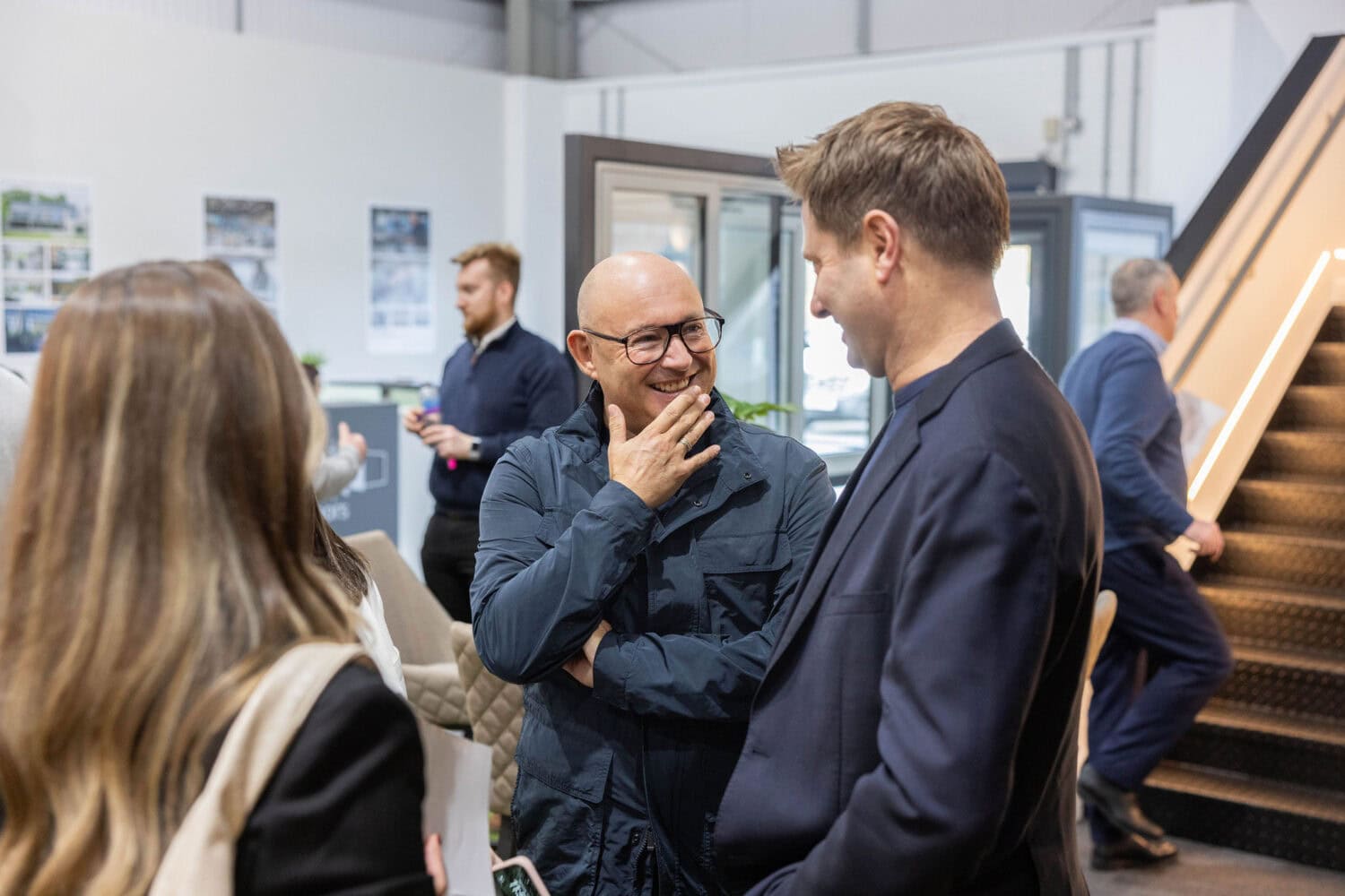 Two men stand talking and smiling at an indoor event, surrounded by others who are conversing. One man is bald and wearing glasses; the other has brown hair. A staircase and large glass roofing are visible in the background.