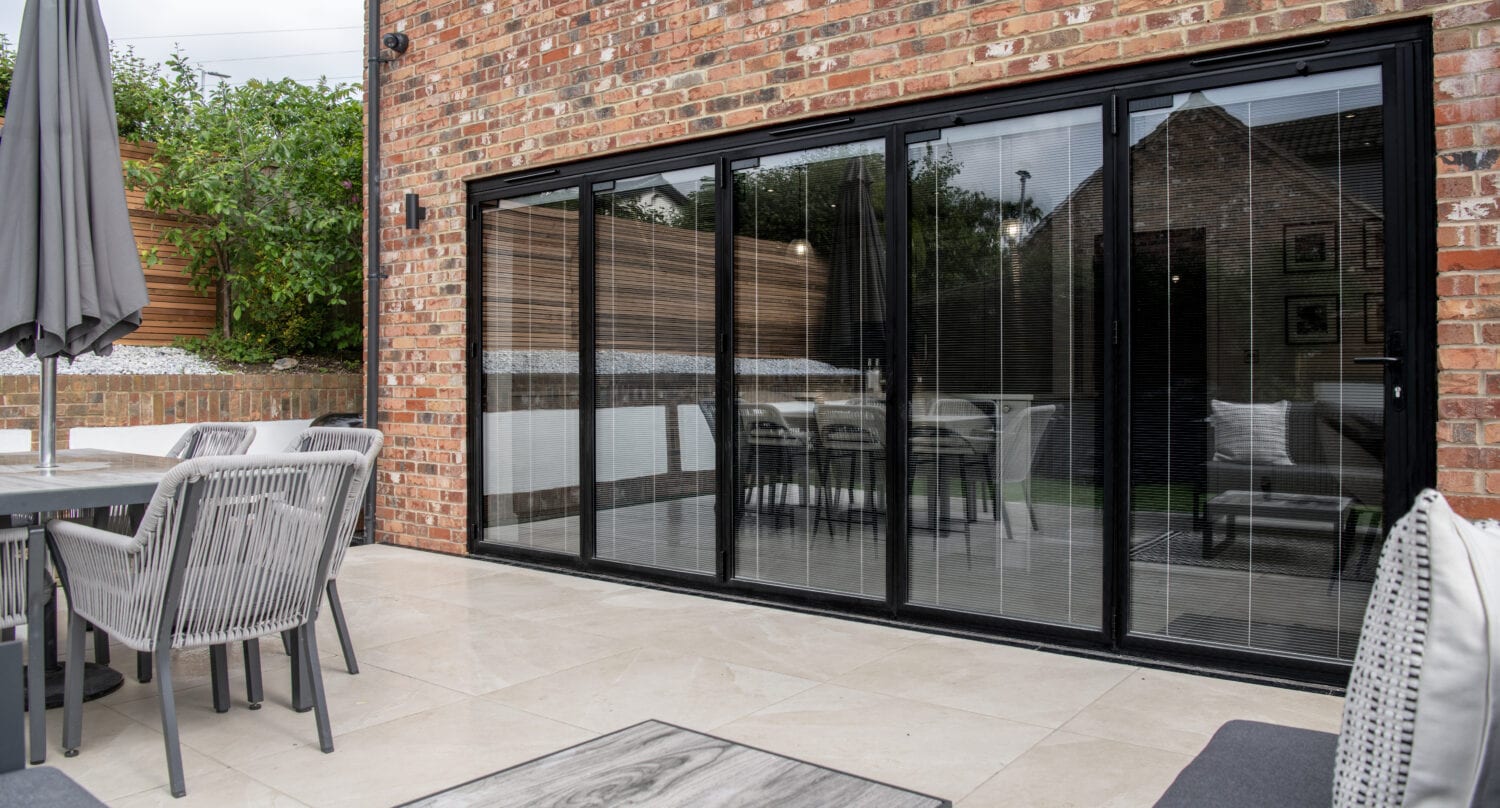A modern patio with outdoor seating, a large table, and an umbrella sits next to a brick house featuring bespoke glass solutions and sliding doors opening into a contemporary dining room.