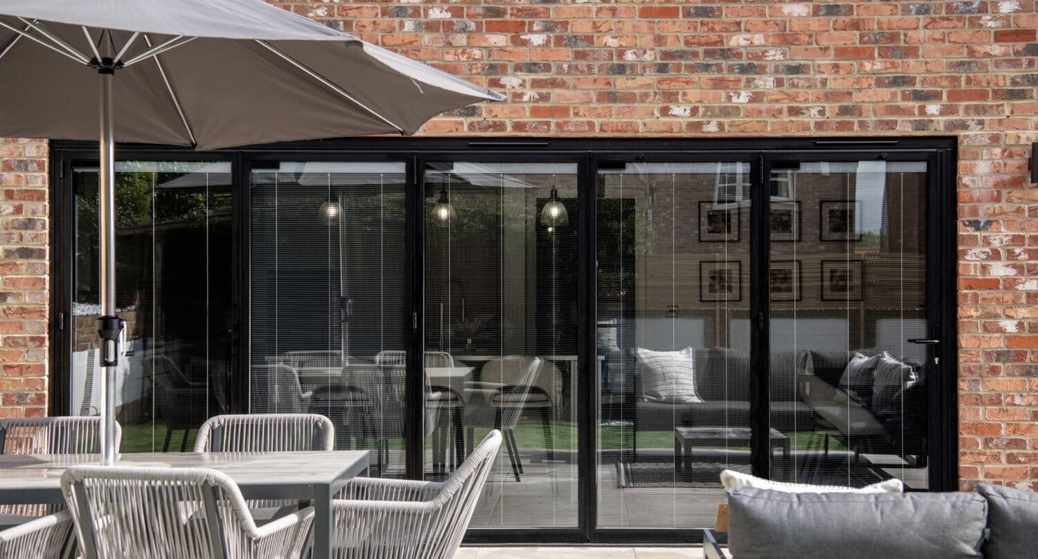A modern patio with a gray outdoor dining set and umbrella sits before large black-framed bespoke glass sliding doors set in a red brick wall, reflecting the garden and interior lights.