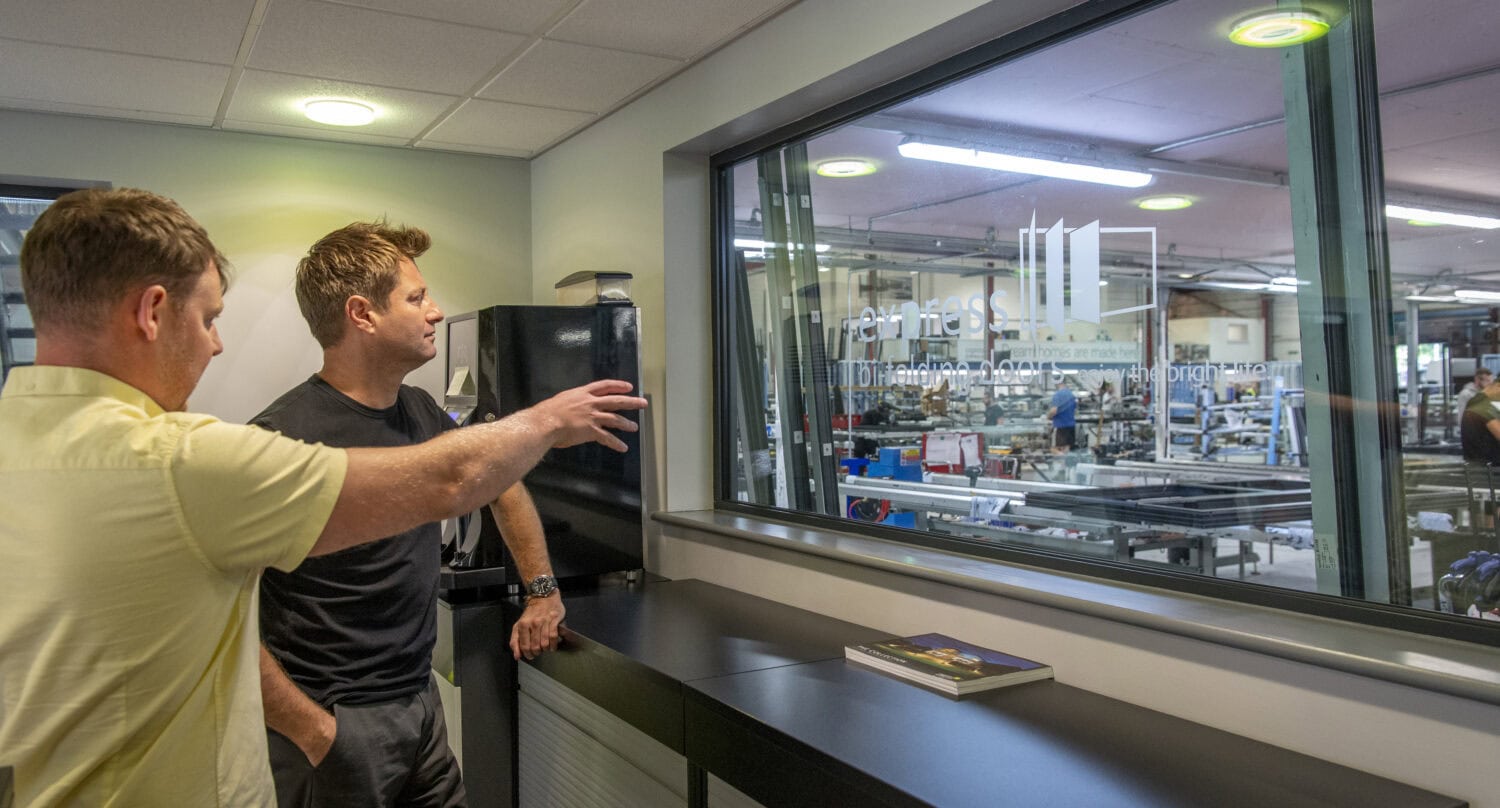 Two men stand indoors by a large window, one gesturing toward a busy factory floor visible through sleek sliding doors, while the other listens attentively. A book and a few items rest on the counter beside them.