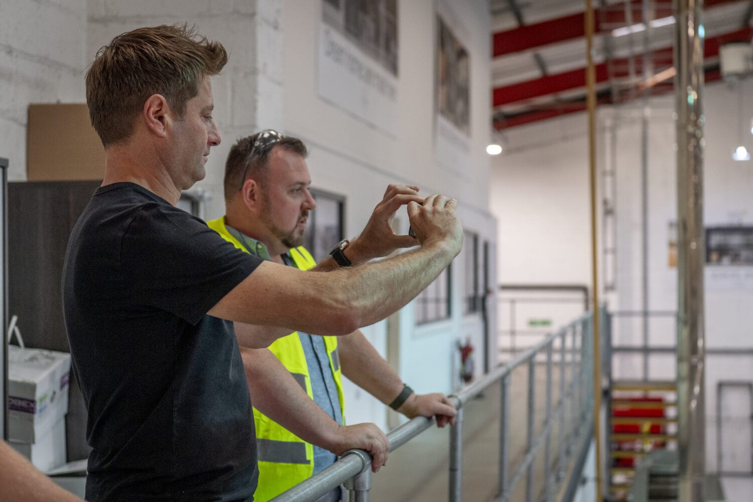 Two men stand on an indoor balcony with bespoke glass solutions; one wears a black shirt and takes a photo with his phone, while the other in a yellow safety vest looks ahead. Industrial equipment and white walls are in the background.
