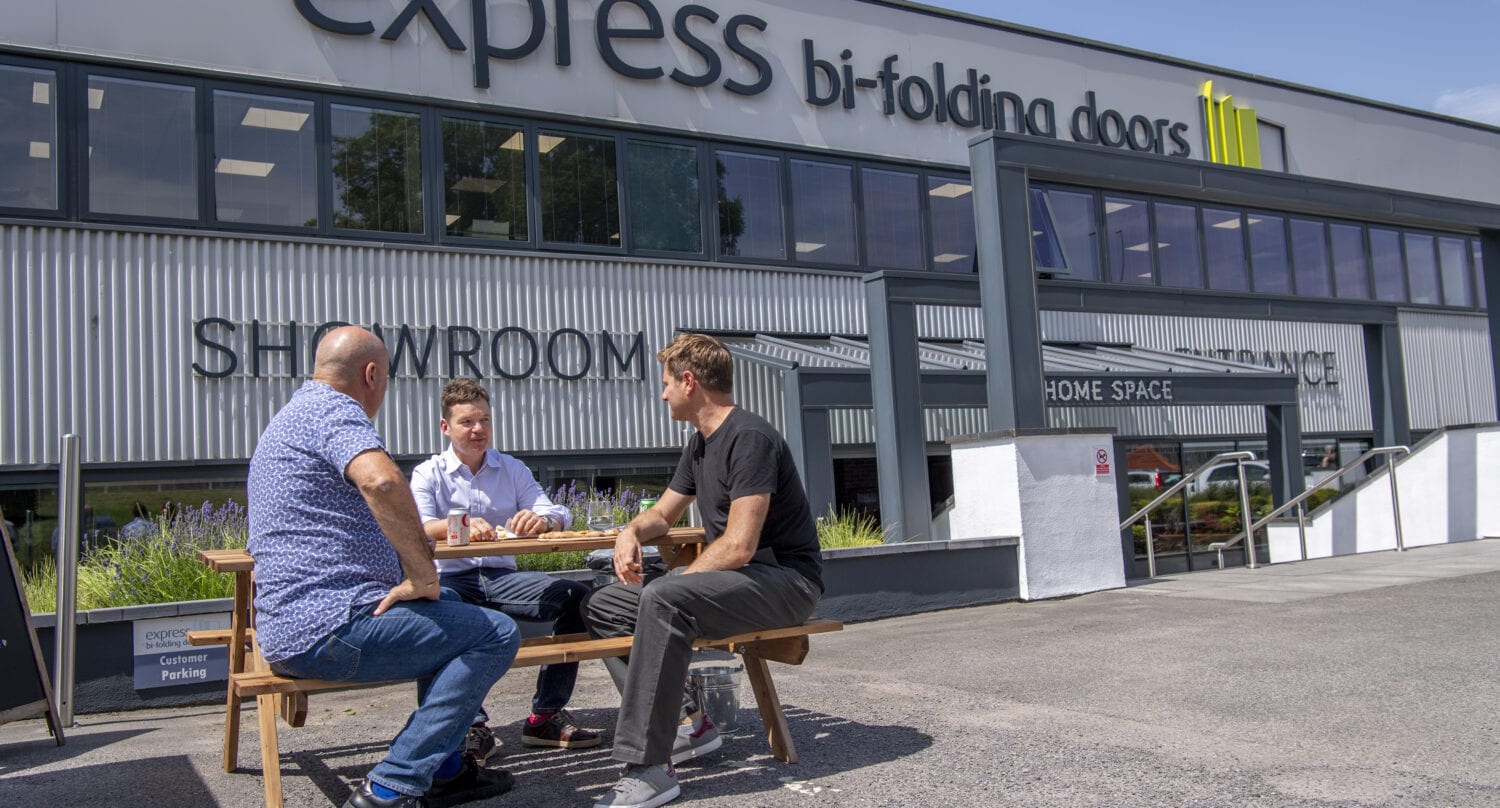 Three men sit and talk around a picnic table outside a large showroom building with a sign that reads express bi-folding doors, known for bespoke glass solutions. The sunny weather adds to the relaxed, casual atmosphere.