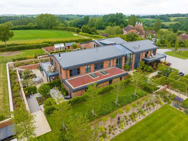 A modern house with brick and glass exterior features sleek bifold doors, surrounded by landscaped gardens, lawns, and trees in a rural area with fields and distant houses visible.