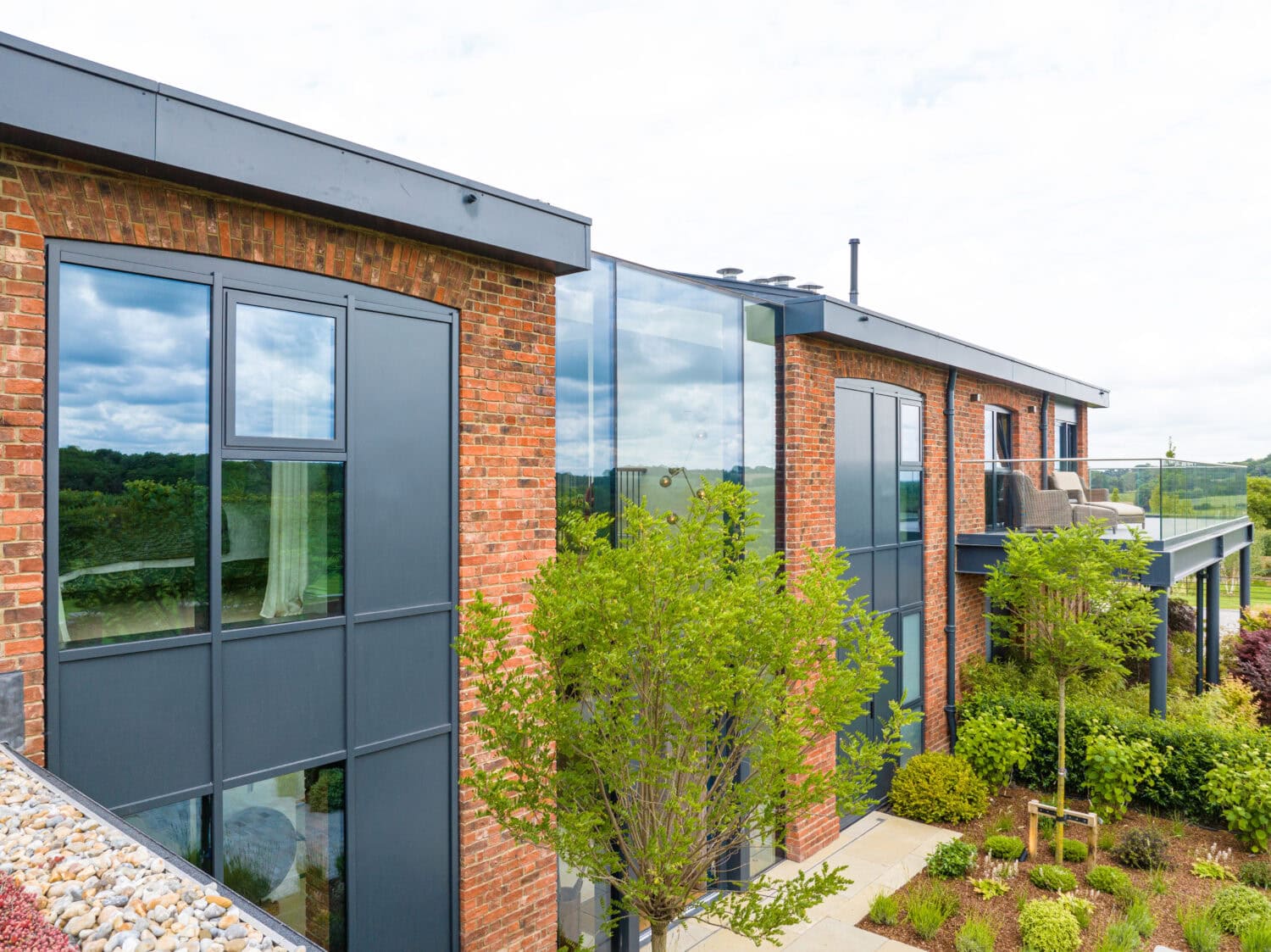 Modern house with large glass windows, brick walls, and sleek sliding doors, surrounded by green trees and landscaped gardens, under a partly cloudy sky.
