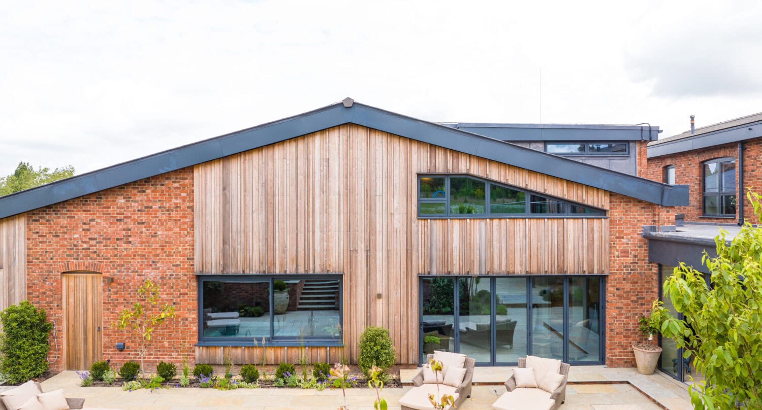 Modern house exterior featuring red brick and wooden paneling, large windows, glass roofing over the patio area with lounge chairs, greenery, and bespoke glass solutions. The sky is partly cloudy.